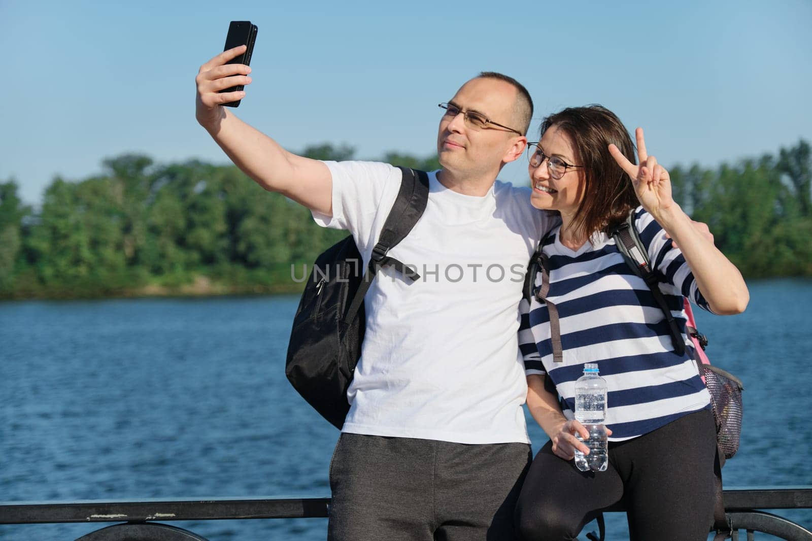Mature happy couple taking selfie photo on phone, people relaxing near river in summer evening park.