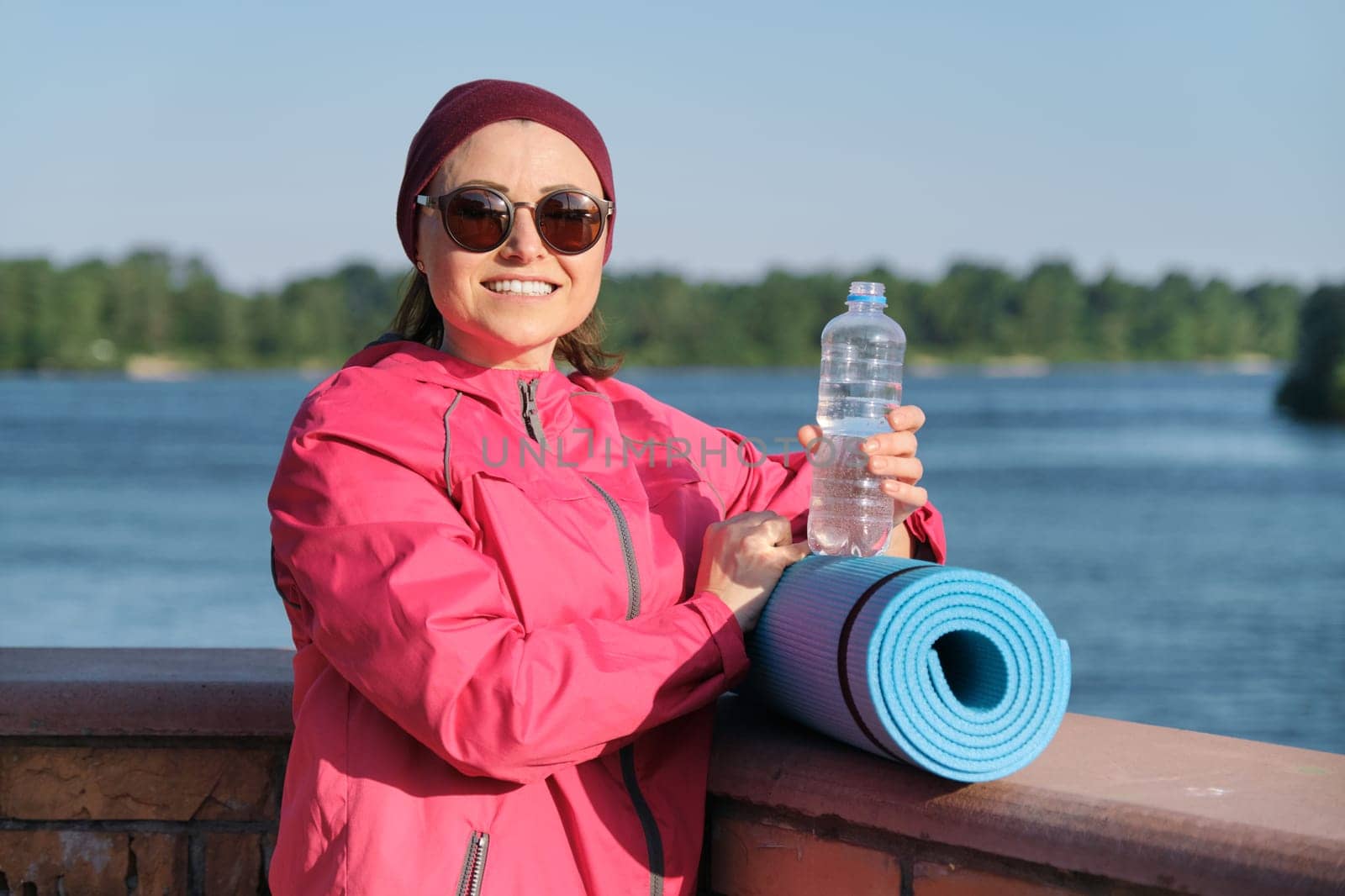 Healthy lifestyle of mature woman, outdoor portrait of an age female in sportswear with yoga mat, drinking water from bottle, background blue sky, river, evening sun