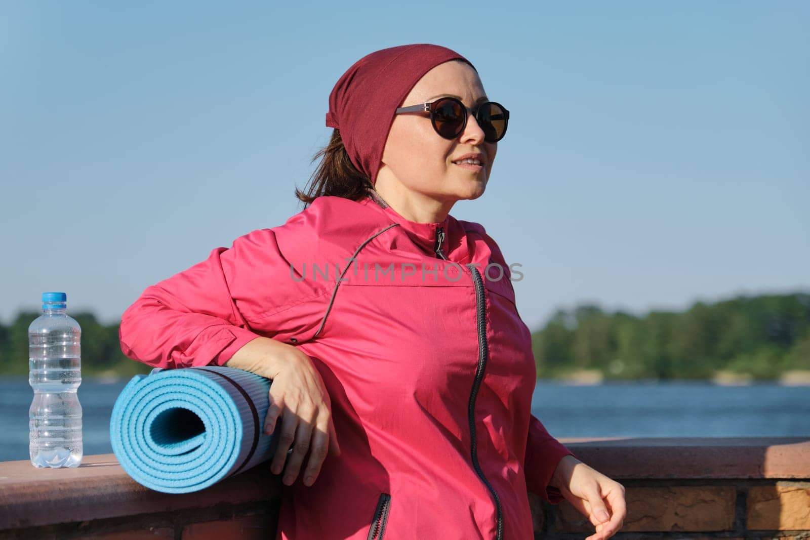 Middle-aged sports woman with yoga mat and bottle of water, an outdoor evening sunny park near the river. Active healthy lifestyle of an age female