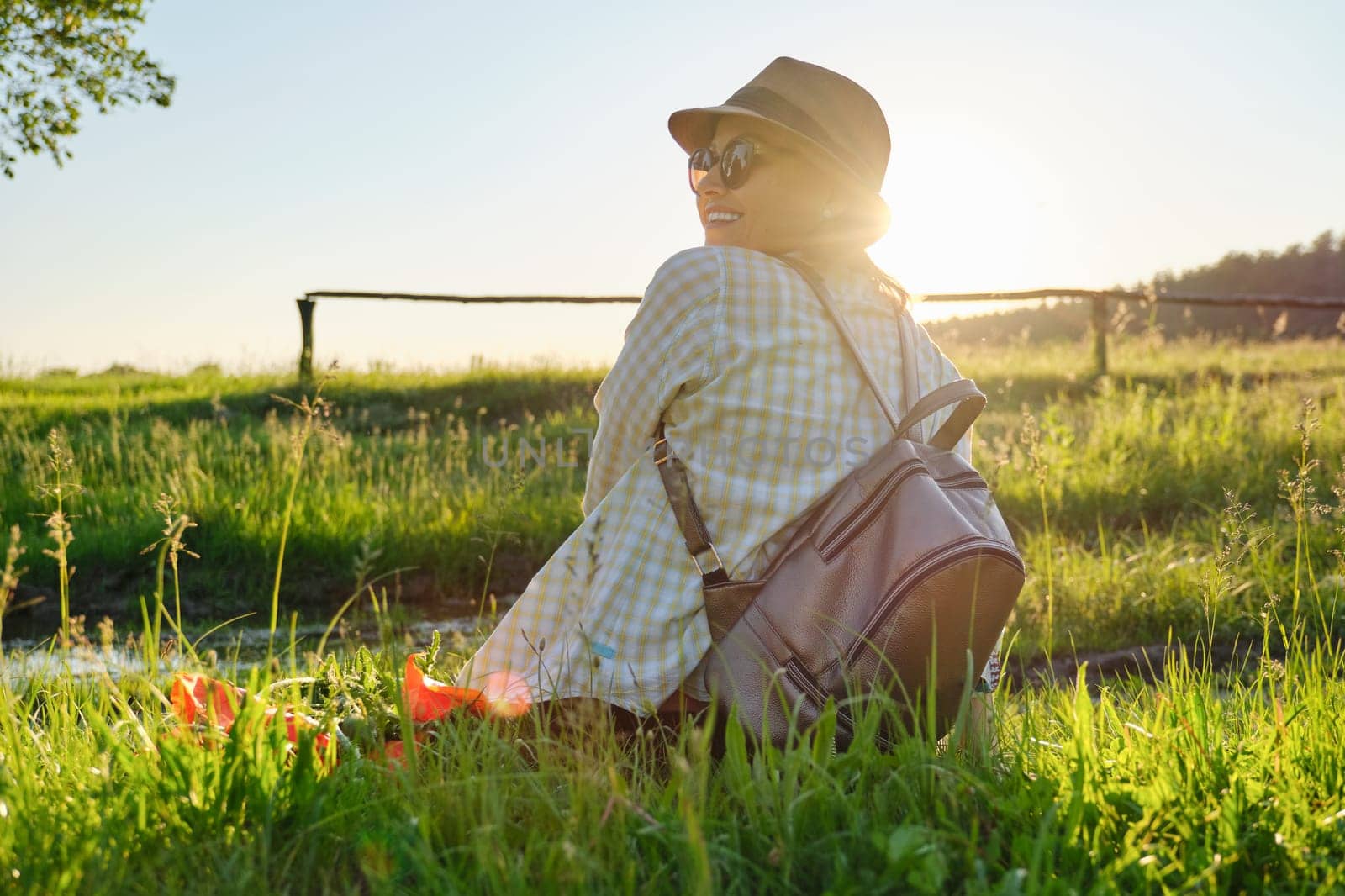 Adult woman in hat with backpack sitting back, summer landscape background, meadow at sunset, golden hour