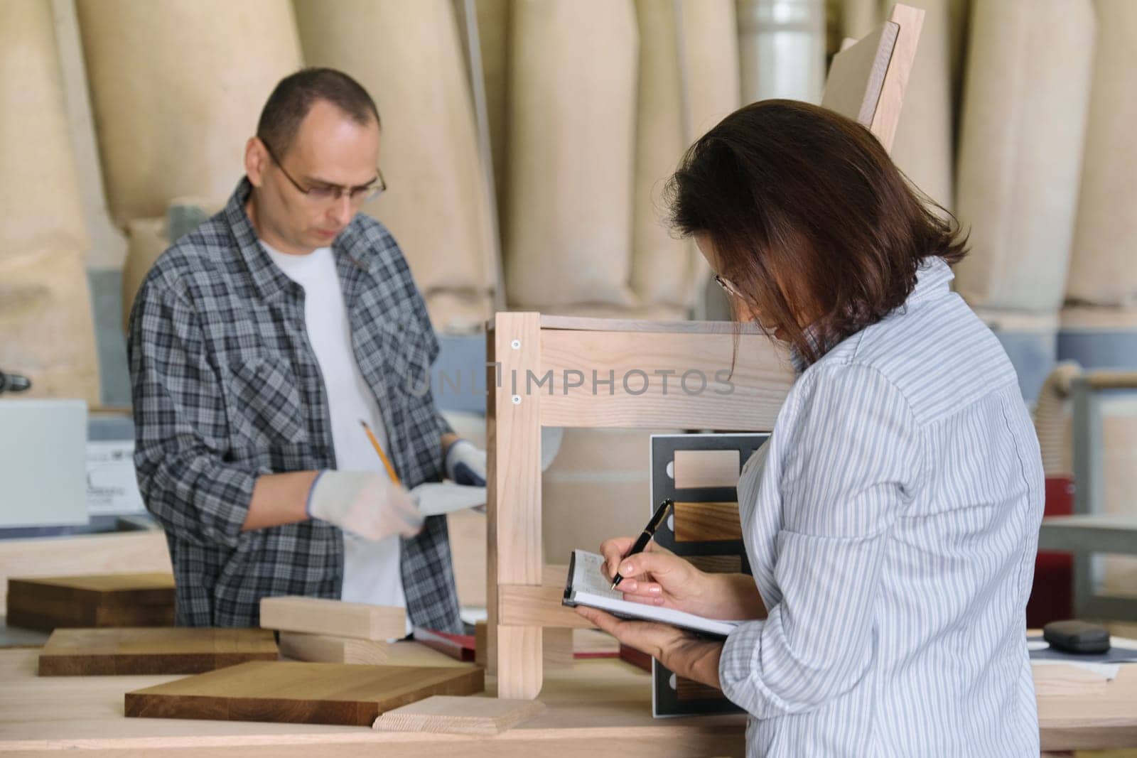 Furniture production master making wooden chair, female designer with wood samples choosing finishing in woodworking workshop.