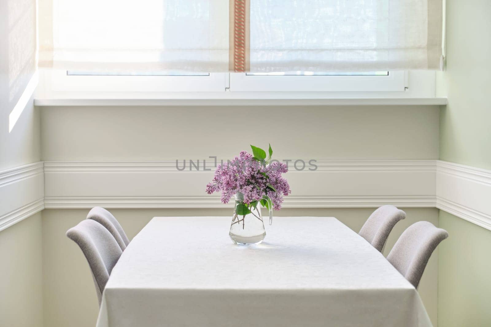 Living room dining area with 4 chairs near window, bouquet of lilac flowers on the table on white tablecloth. Home interior decor