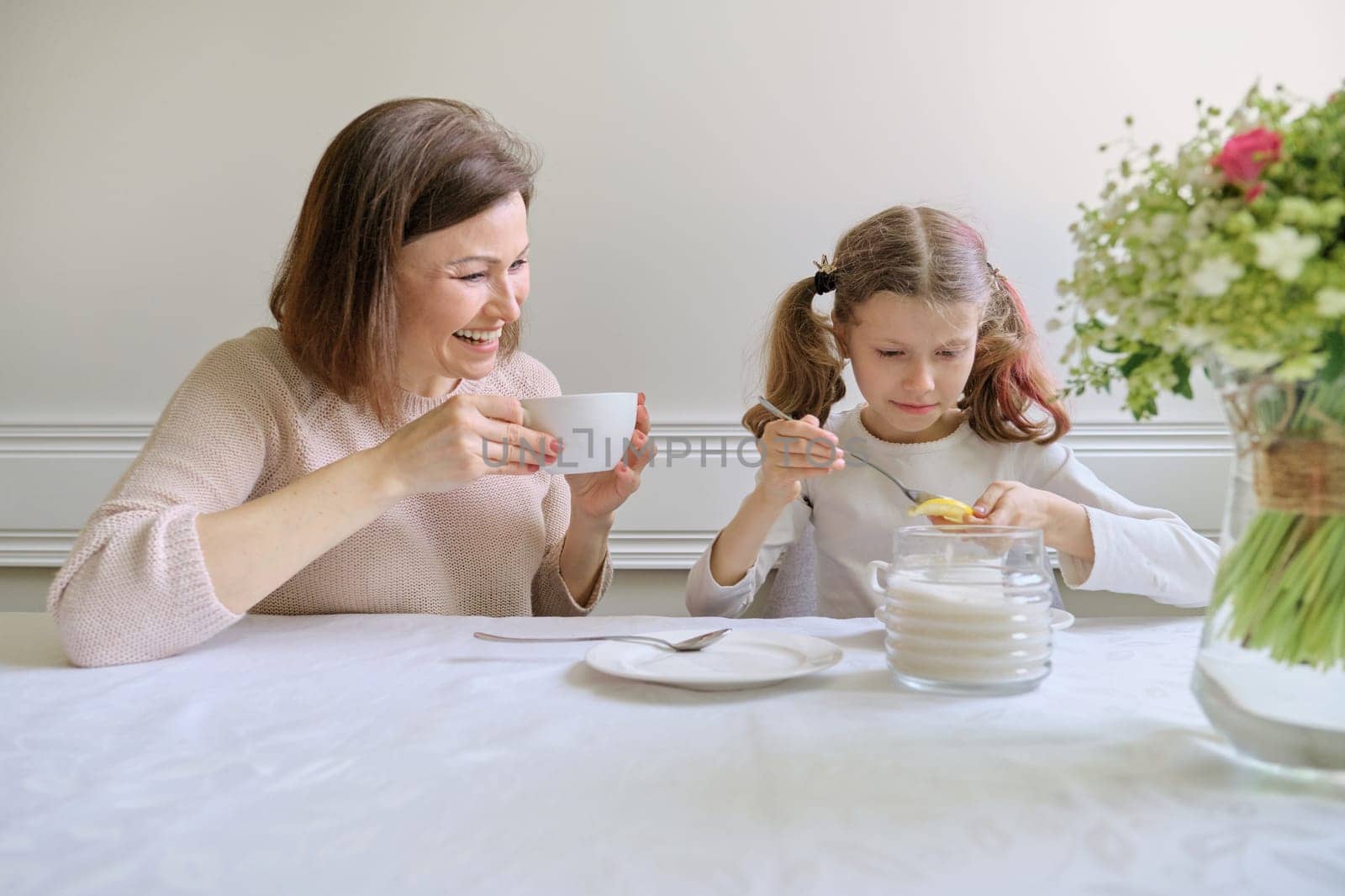 Laughing mother and little daughter, sitting at home at the table drinking from cups and eating lemon. Parent and child having fun and talking