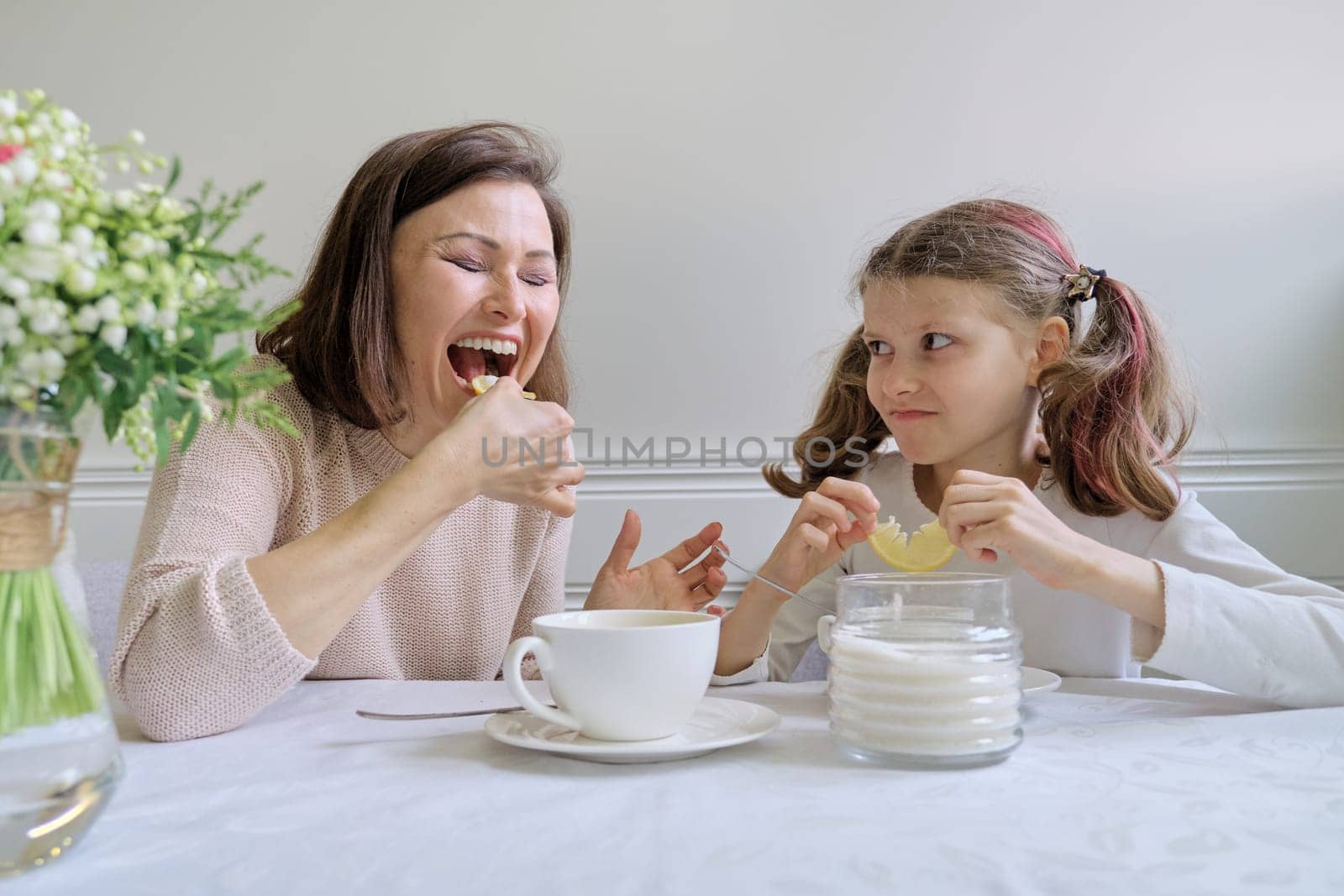 Laughing mother and little daughter, sitting at home at the table drinking from cups and eating lemon. Parent and child having fun and talking