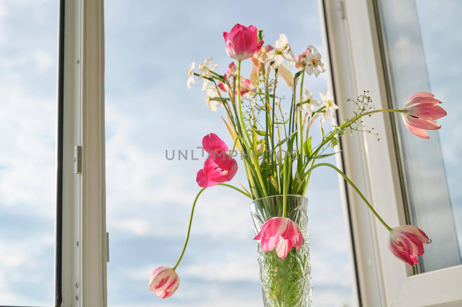 Bouquet of spring flowers tulips and white daffodils in vase on the window, background evening sunset sky with clouds
