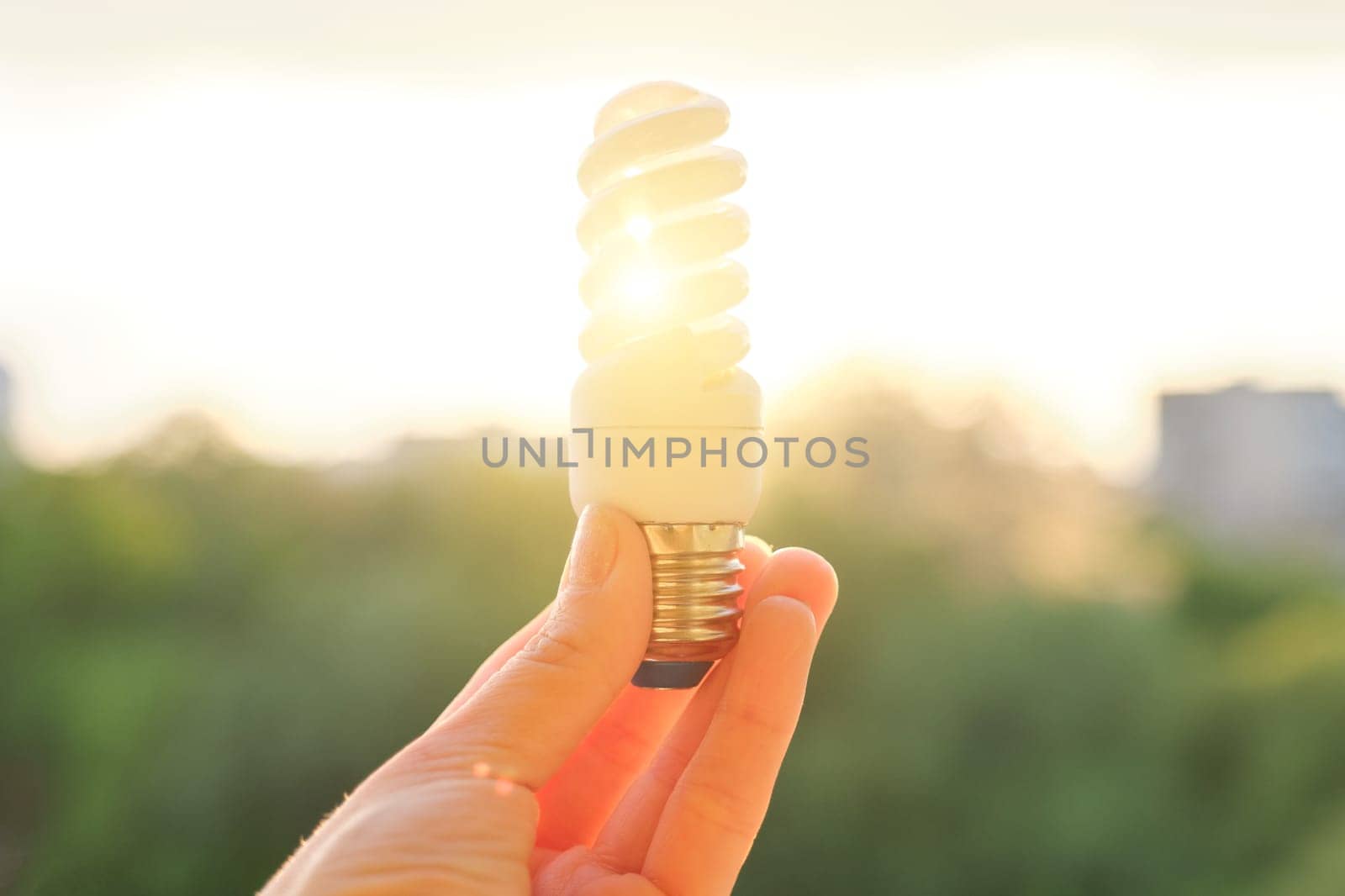 Energy saving fluorescent light bulb, hand holding lamp, evening sunset sky background.