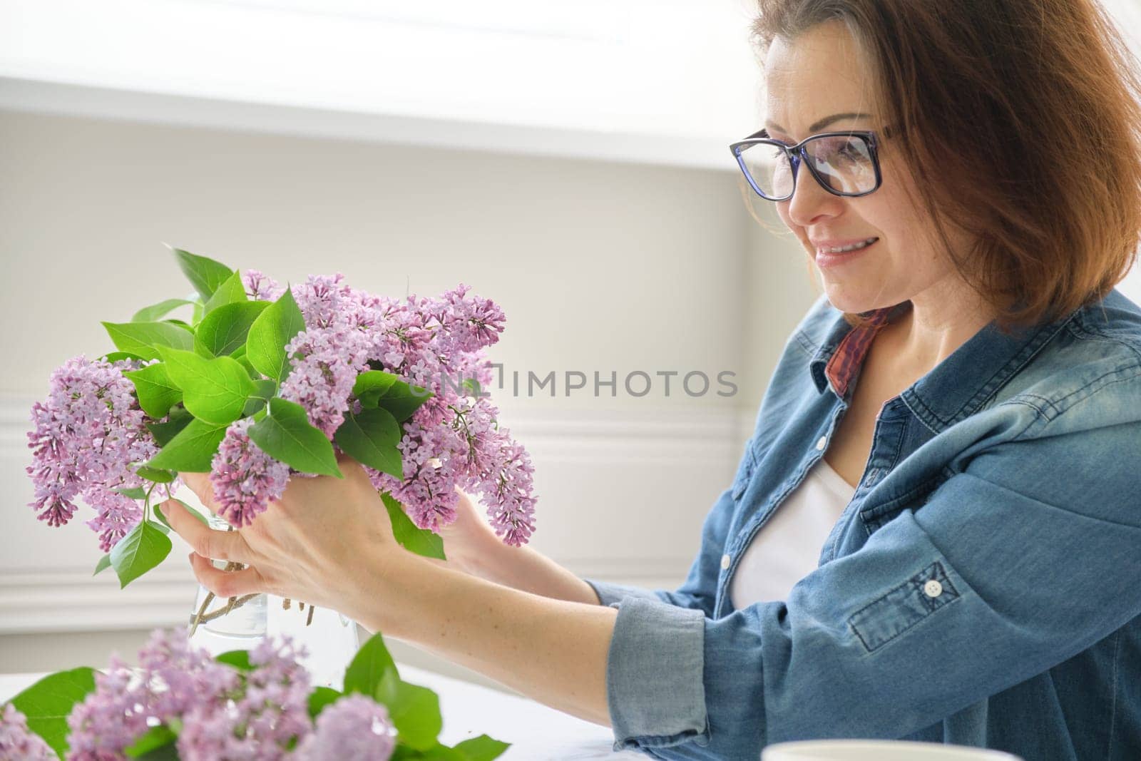Middle-aged woman making bouquet of lilac branches, close-up female arranging flowers bunch in vase by VH-studio