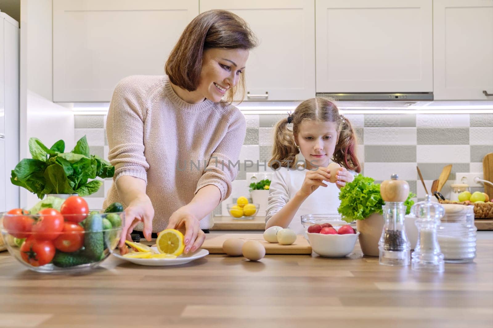 Smiling mother and daughter 8, 9 years old cooking together in kitchen vegetable salad. Healthy home food, communication parent and child. Woman cutting lemon, girl shelling chicken eggs.