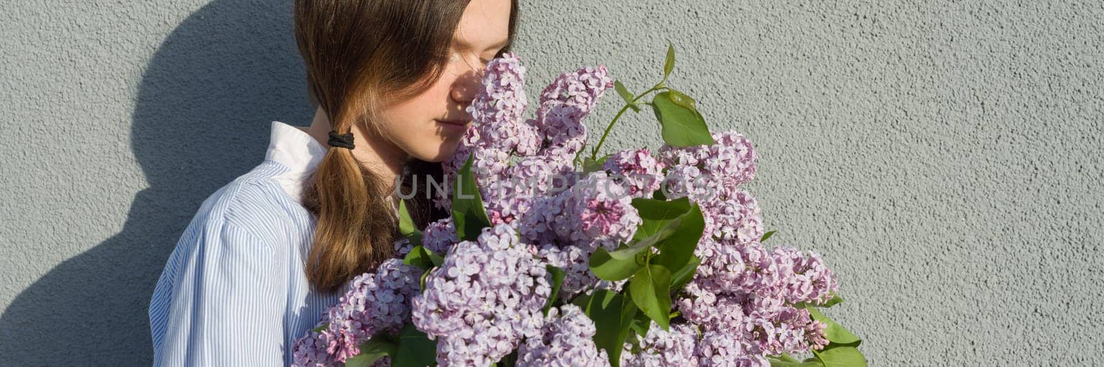 Young teen girl with bouquet of lilac near gray wall outdoor background, panoramic banner, girl in profile, copy space