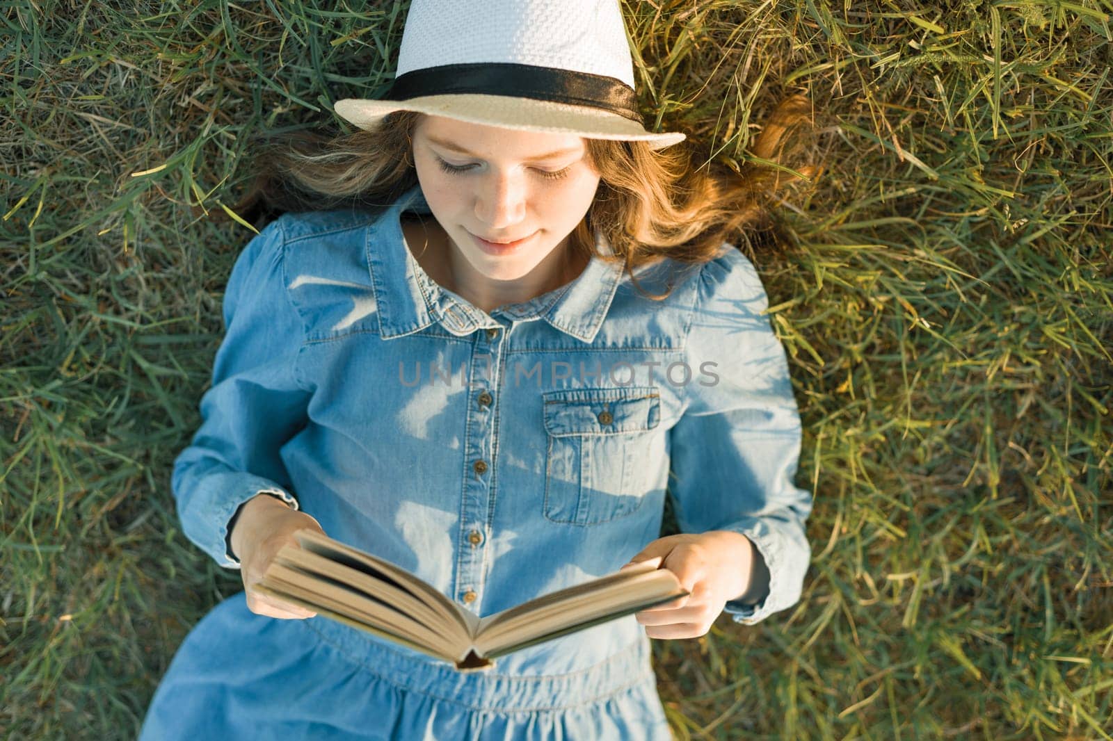 Girl in dress and hat lies on green grass reading book. View from above.
