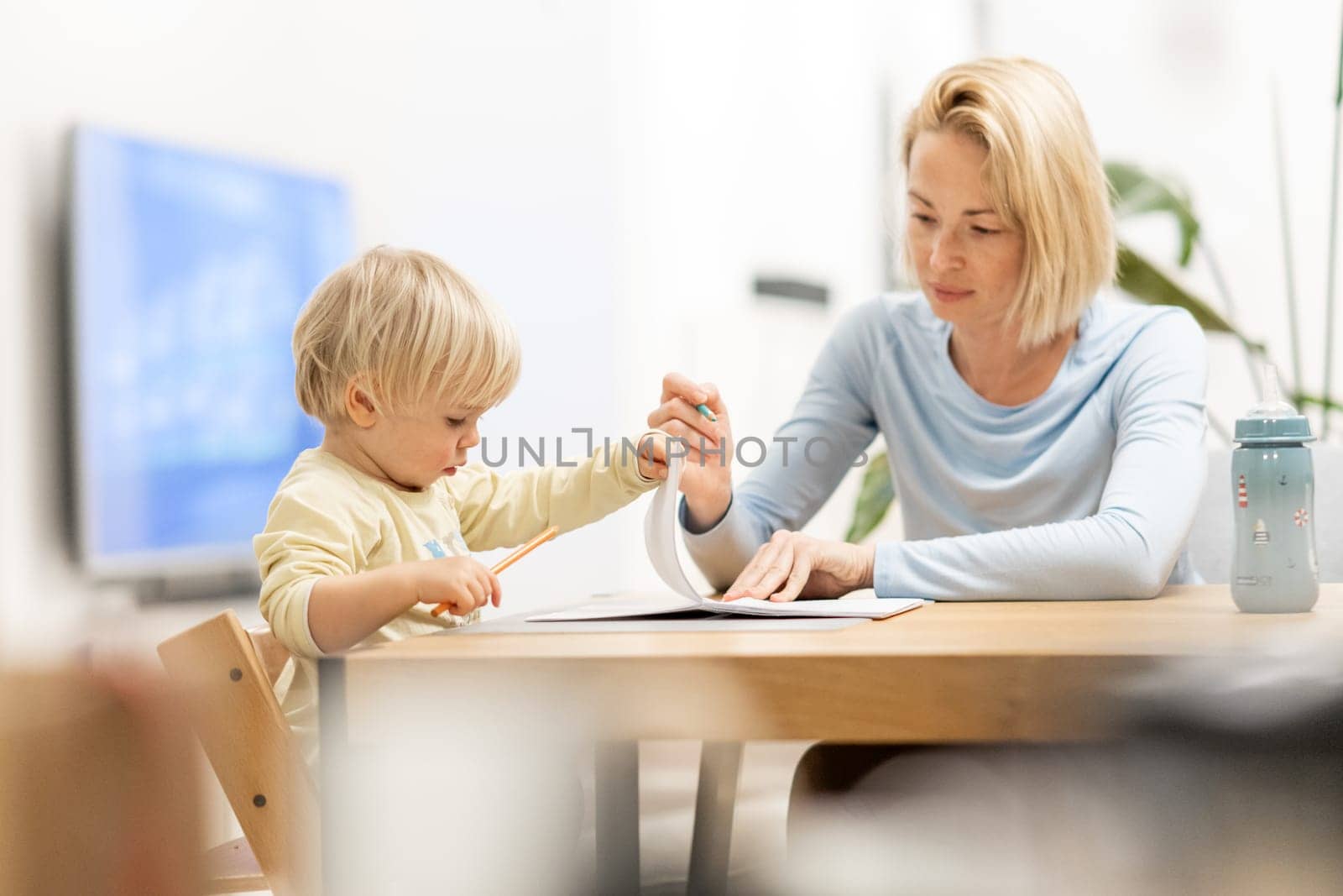 Caring young Caucasian mother and small son drawing painting in notebook at home together. Loving mom or nanny having fun learning and playing with her little 1,5 year old infant baby boy child