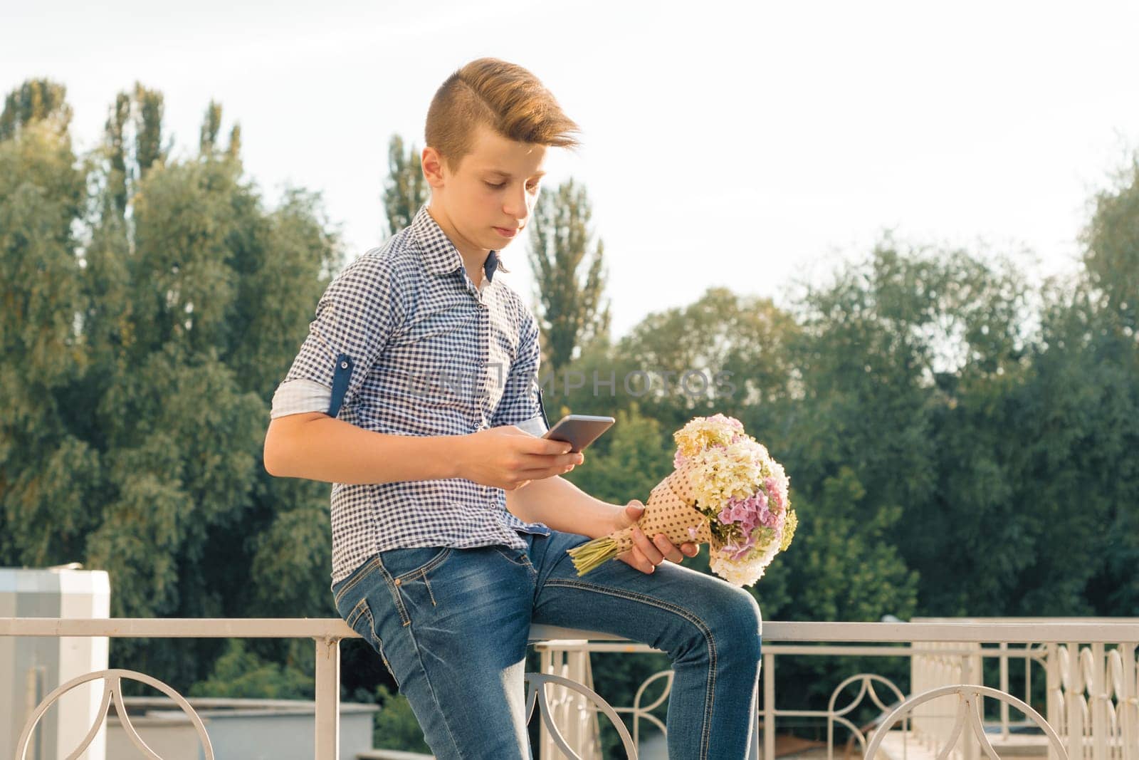 Outdoor portrait of teenage boy with bouquet of flowers, reading text on smartphone, urban background.