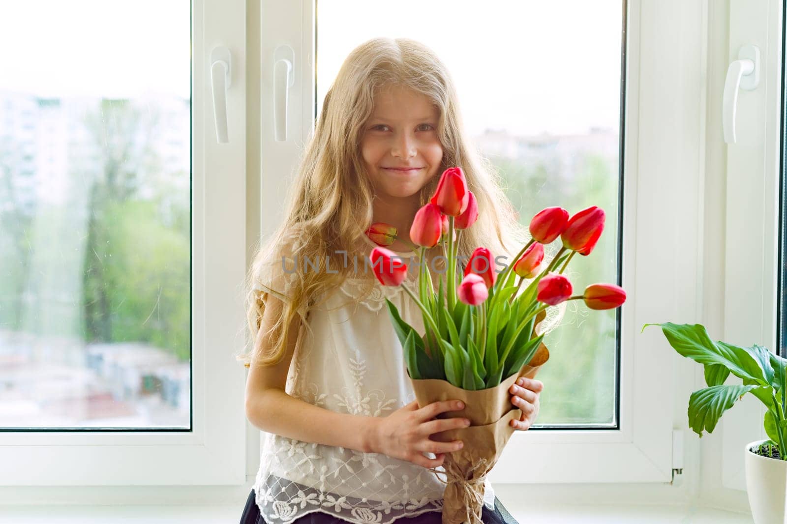 Little beautiful child girl with bouquet of red tulips flowers at home near the window, present for mother day. Gift, surprise, spring family holiday. Mothers Day