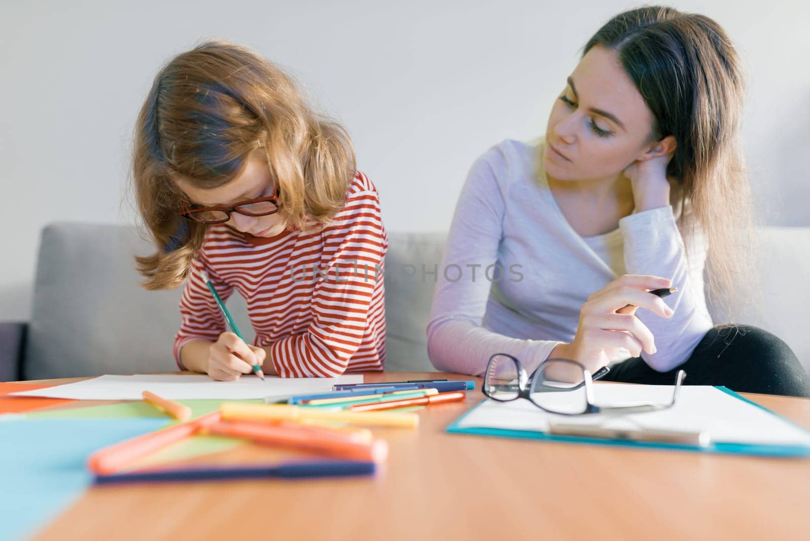 Young female teacher giving private lesson to child, little girl sitting at her desk writing in notebook.