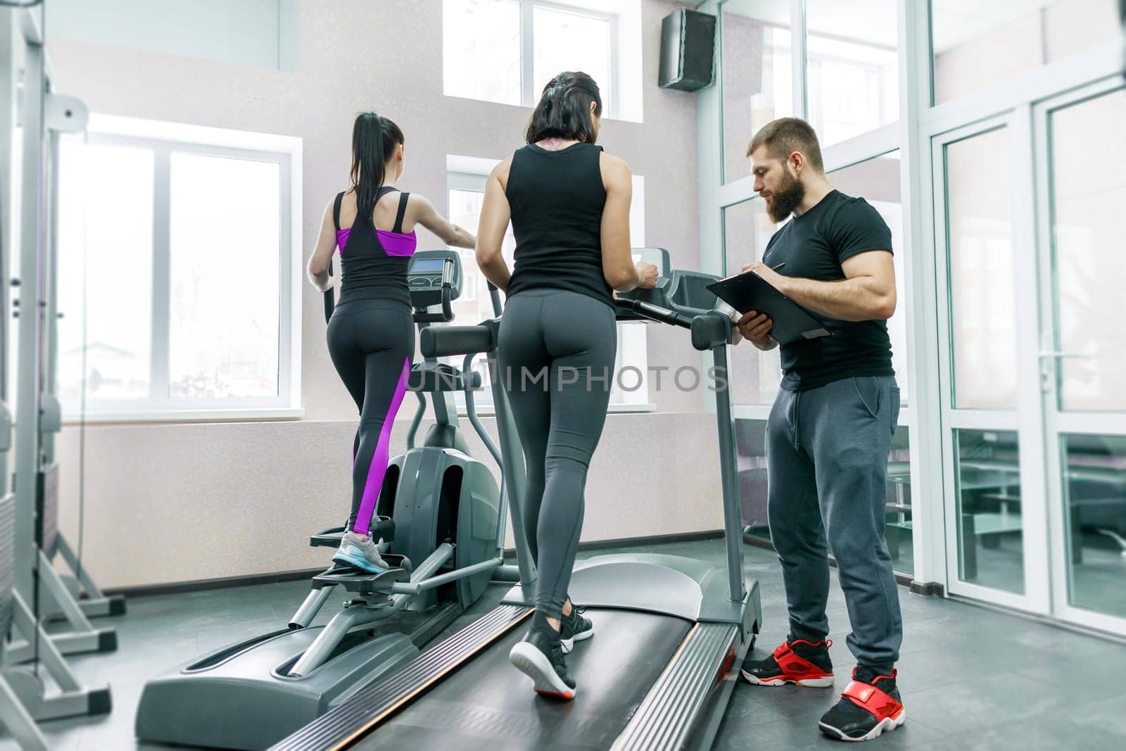 Young smiling fitness women with personal trainer an adult athletic man on treadmill in the gym. Sport, teamwork, training, healthy lifestyle concept.