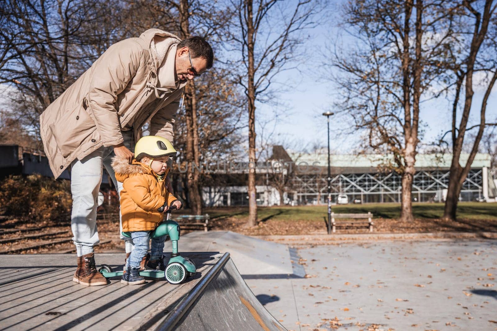 Father supervises his fearless small toddler boy while riding baby scooter outdoors in urban skate park. Child wearing yellow protective helmet by kasto