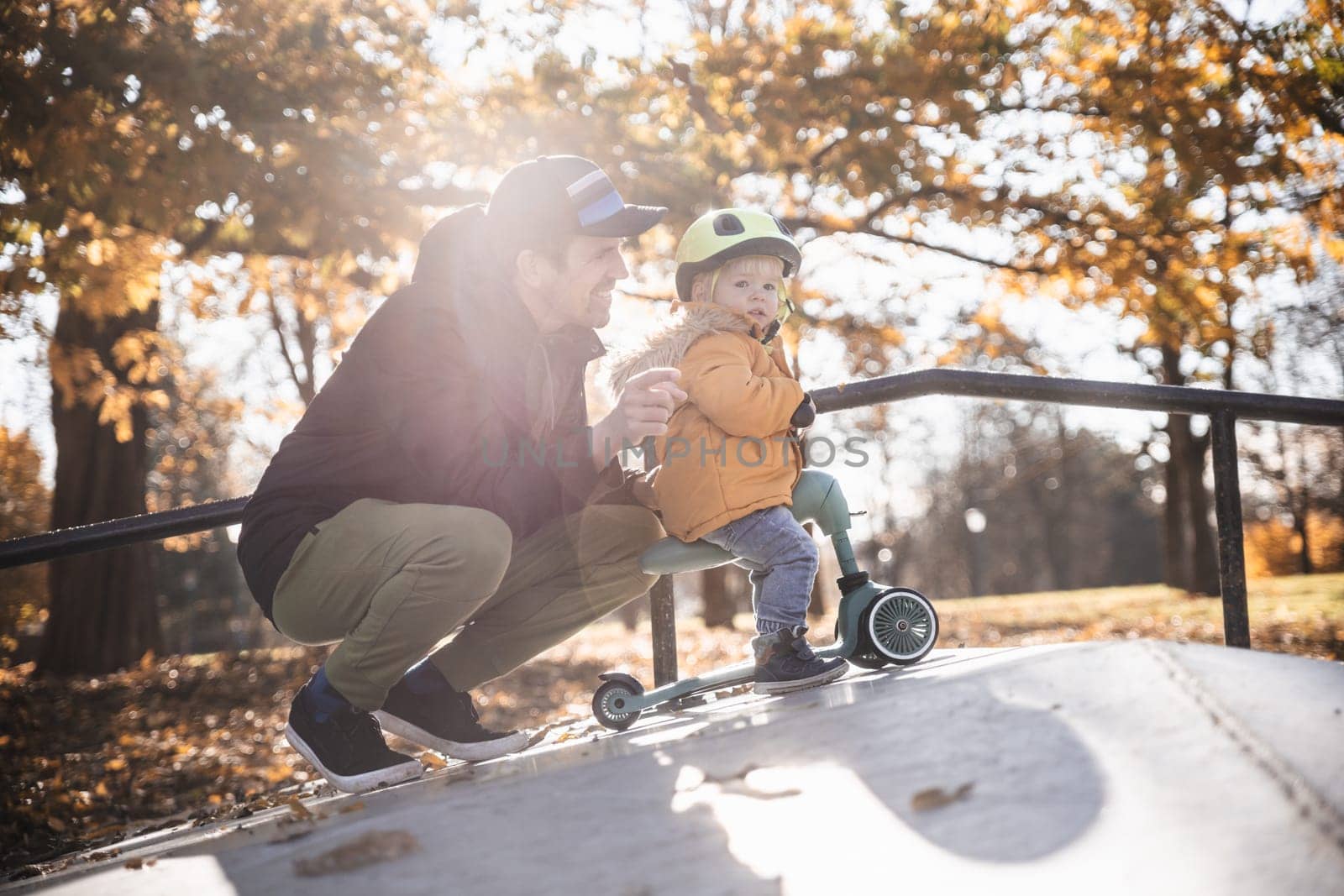 Father supervises his fearless small toddler boy while riding baby scooter outdoors in urban skate park. Child wearing yellow protective helmet by kasto