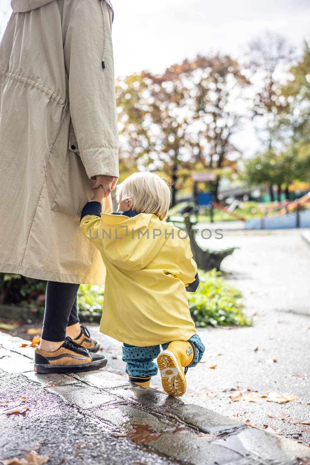 Small bond infant boy wearing yellow rubber boots and yellow waterproof raincoat walking in puddles on a overcast rainy day holding her mother's hand. Mom with small child in rain outdoors