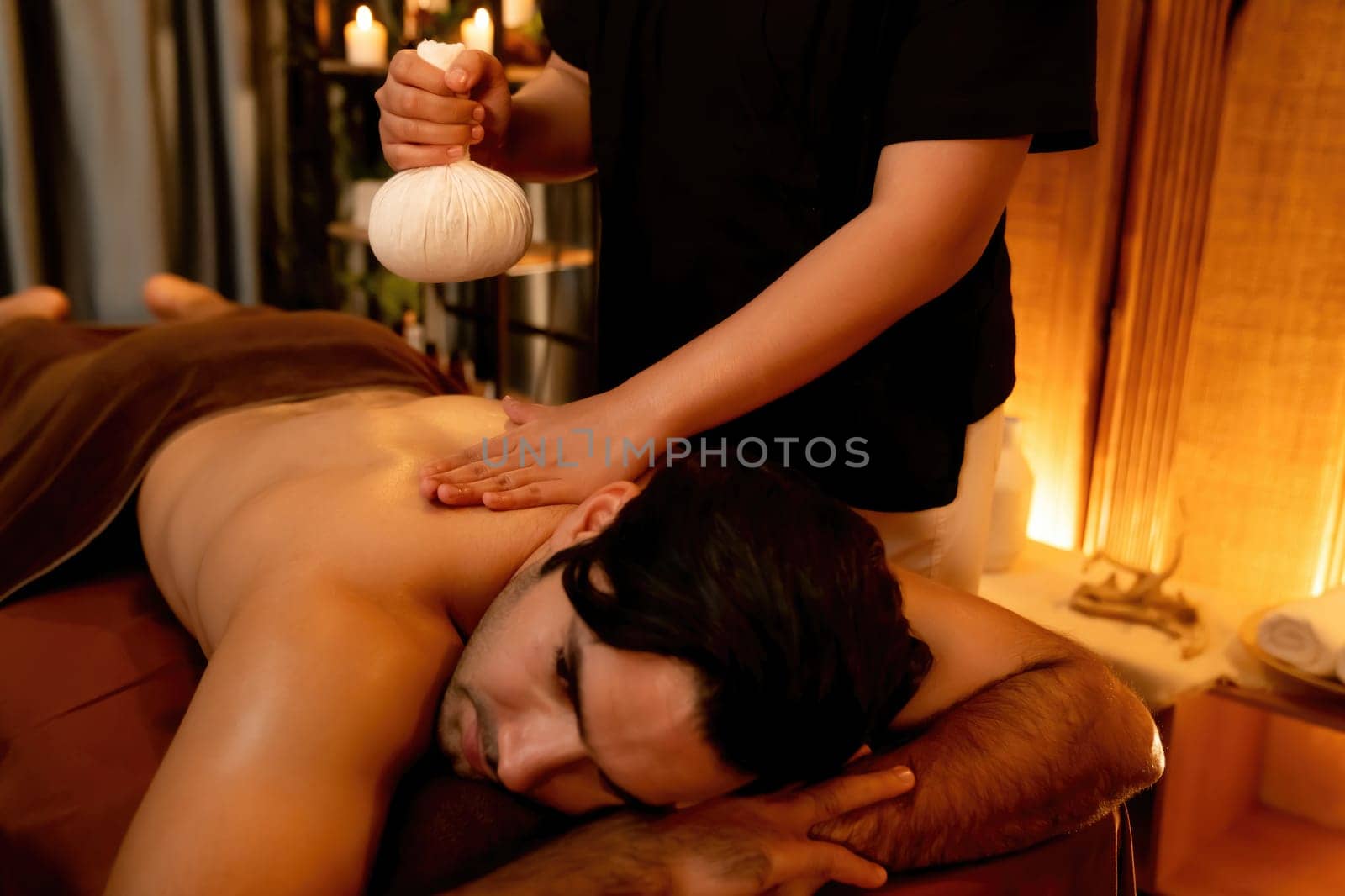 Caucasian man customer enjoying relaxing anti-stress spa massage and pampering with beauty skin recreation leisure in warm candle lighting ambient salon spa at luxury resort or hotel. Quiescent