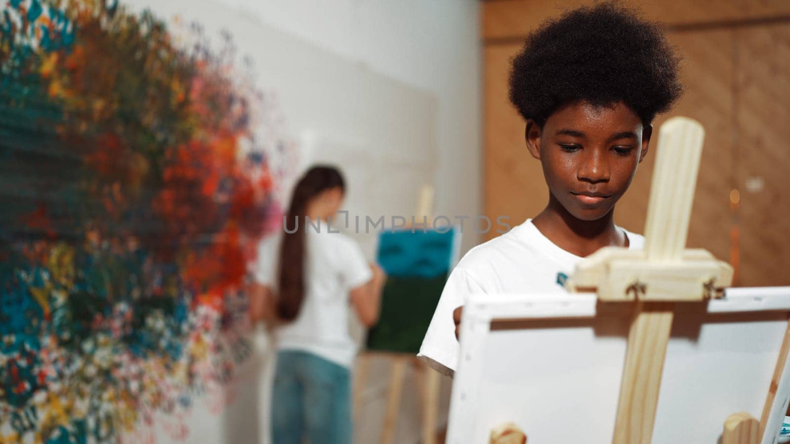 Smart african boy painting canvas while cute girl drawing behind. in art lesson. Highschool student create artwork by express creativity idea while standing at colorful stained wall. Edification.