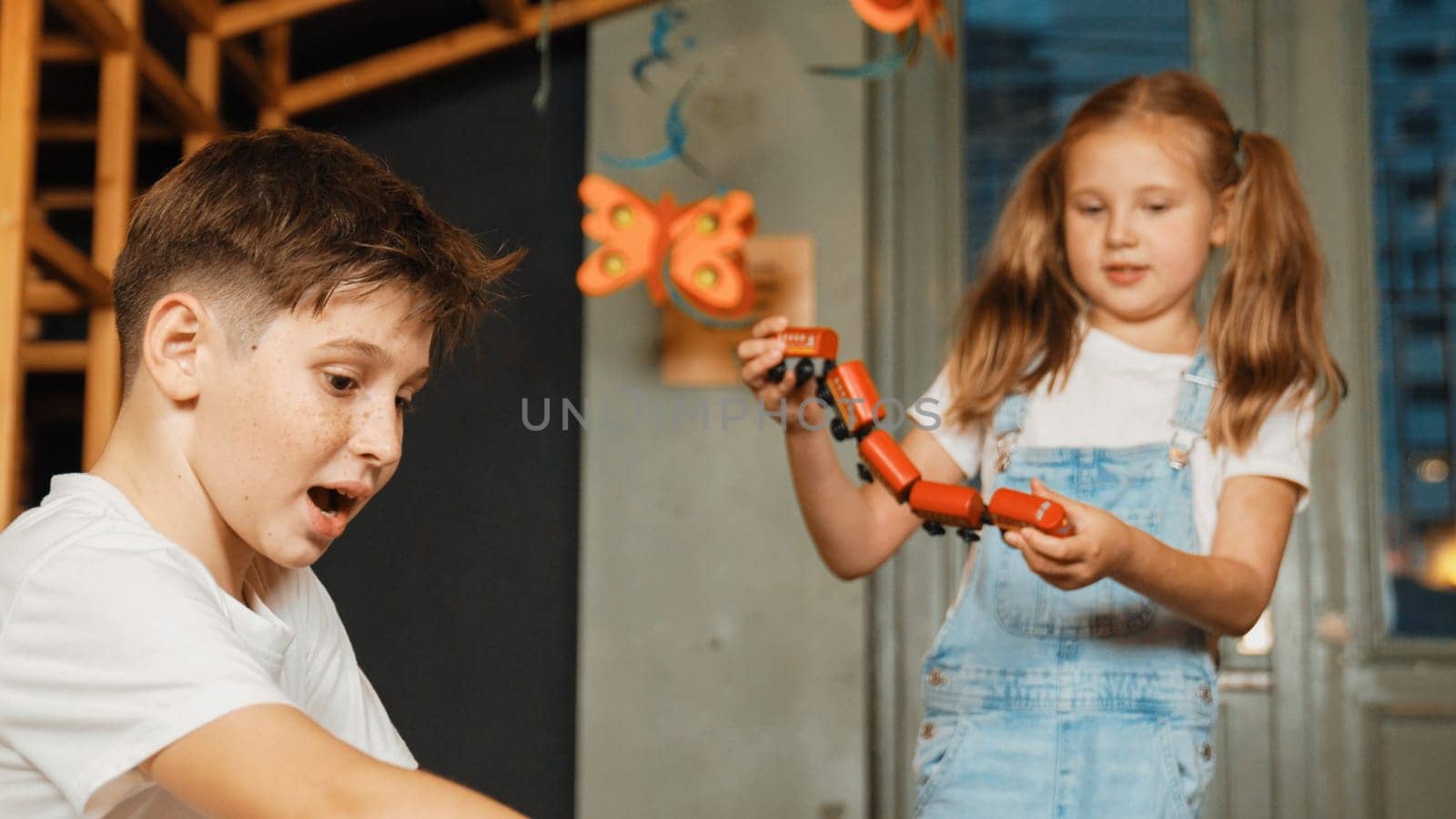 Girl playing wooden train while diverse student sitting at play room. Boy looking at lovely cute student hold toy while his friend talking about lesson in break time. Creative activity. Erudition.