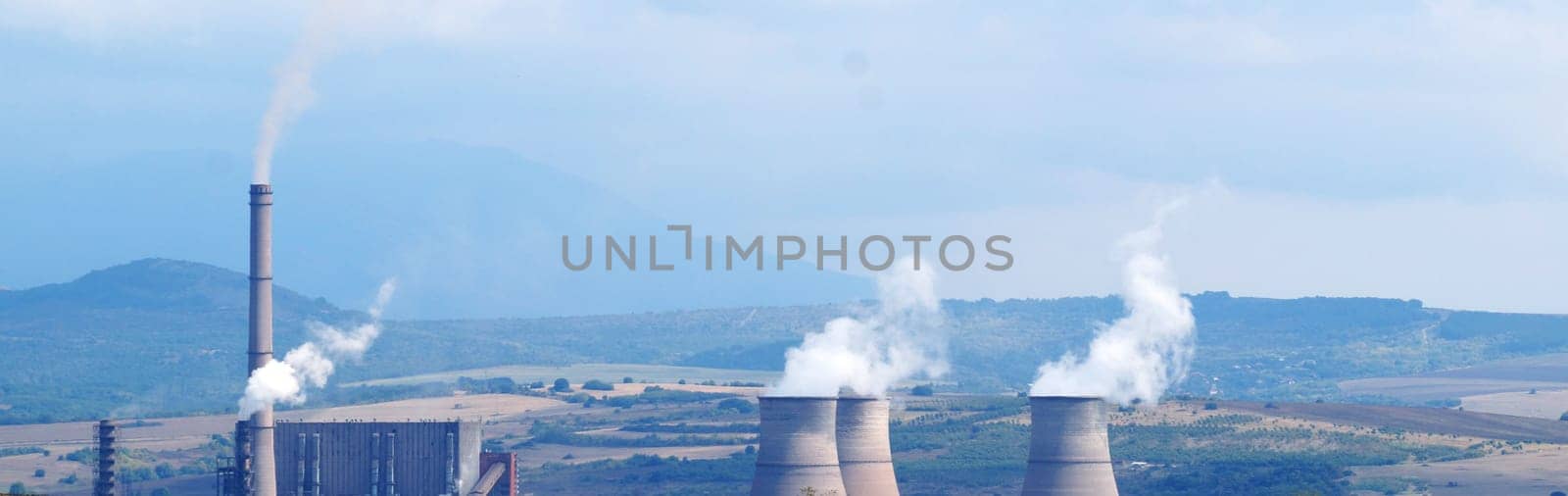 Smoking chimneys of a thermal power plant against the backdrop of fields and mountains, panoramic photo in blue tones