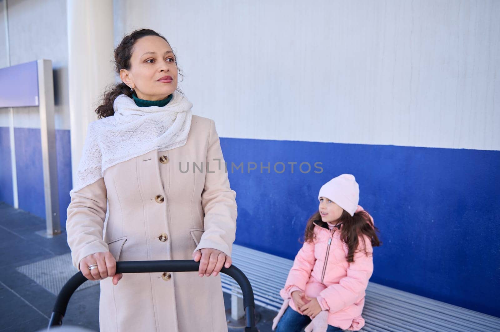 Young woman, mother pushing baby pram, standing near her daughter on the railway station platform, waiting for train by artgf