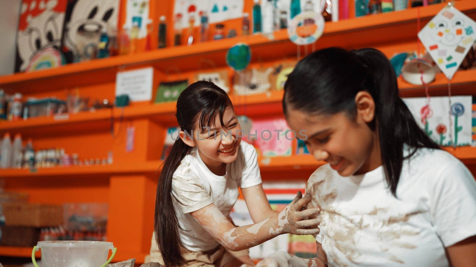 Happy multicultural girl and friend put the clay on the each other muddy shirt at art lesson. Diverse highschool student playing with clay at pottery workshop. Creative activity concept. Edification.