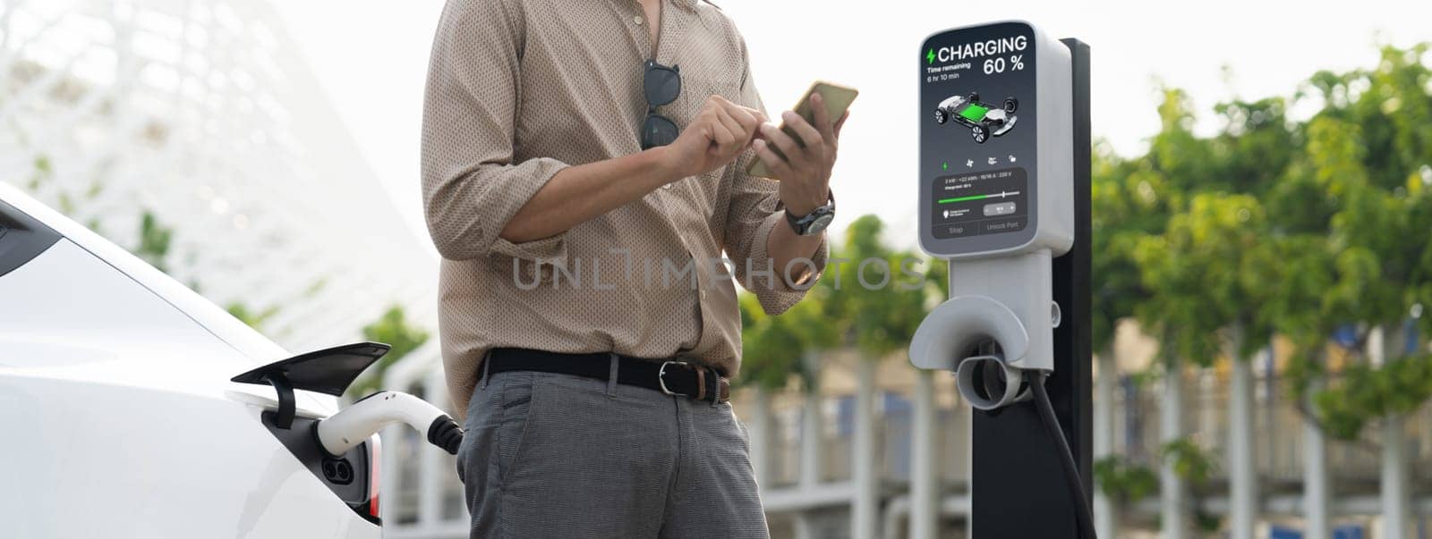 Young man use smartphone to pay for electricity at public EV car charging station green city park. Modern environmental and sustainable urban lifestyle with EV vehicle. Panorama Expedient