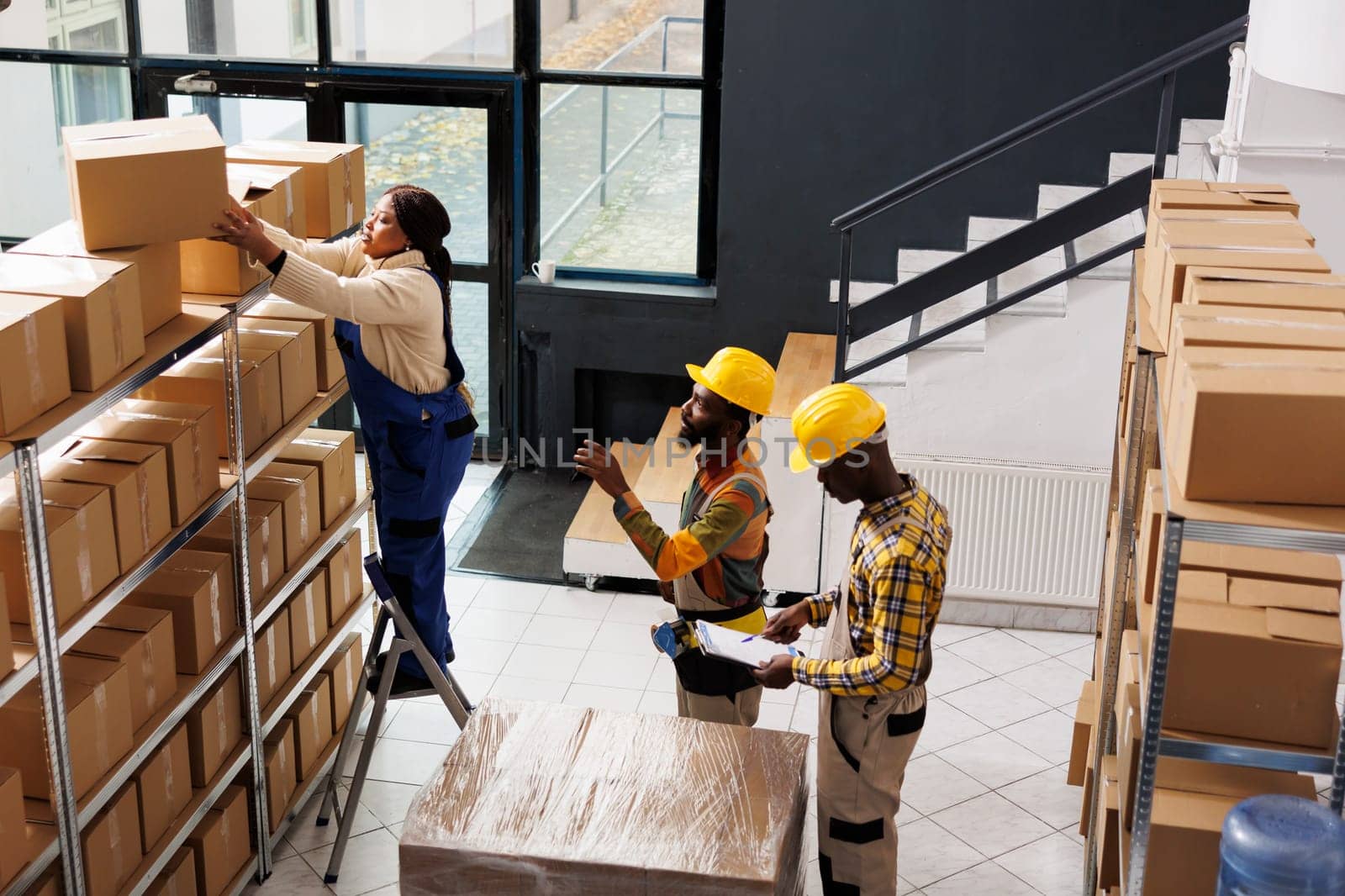 African american post office employee standing on ladder and taking parcel from shelf. All black package handlers team inspecting order and preparing cardboard box for shipment top view