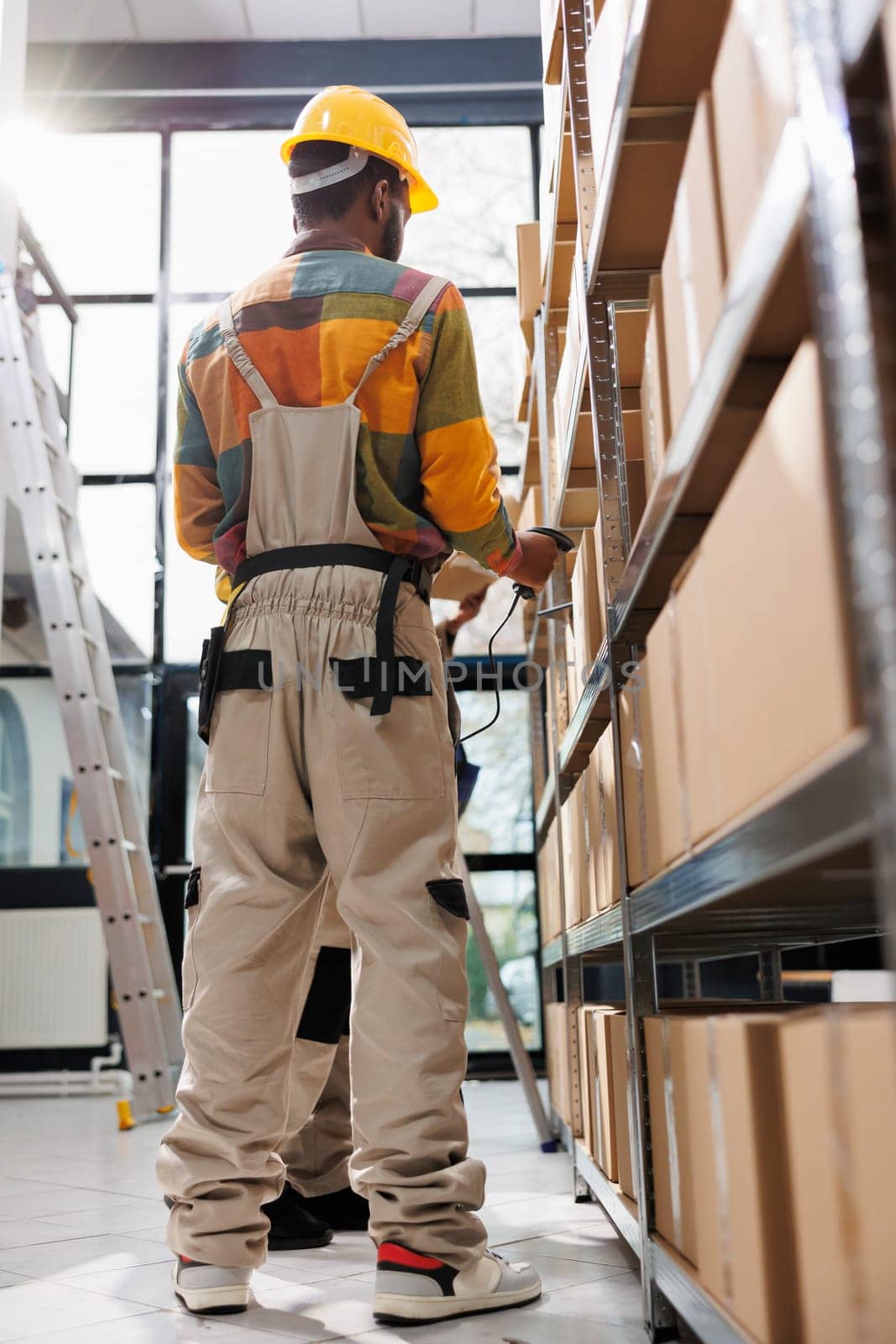 Warehouse employees managing products storing using barcode scanner. African american industrial storehouse employees wearing safety workwear standing near cardboard boxes shelf and scanning parcel