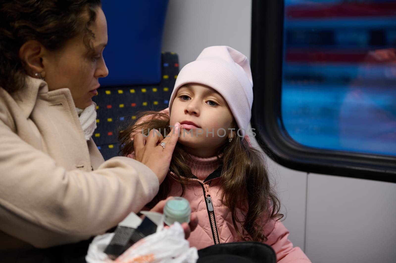Portrait of a young woman applying moisturizing lip balm on her daughter lips, taking care of skin in the winter time. The problem of dry air in urban transport during cold winter time