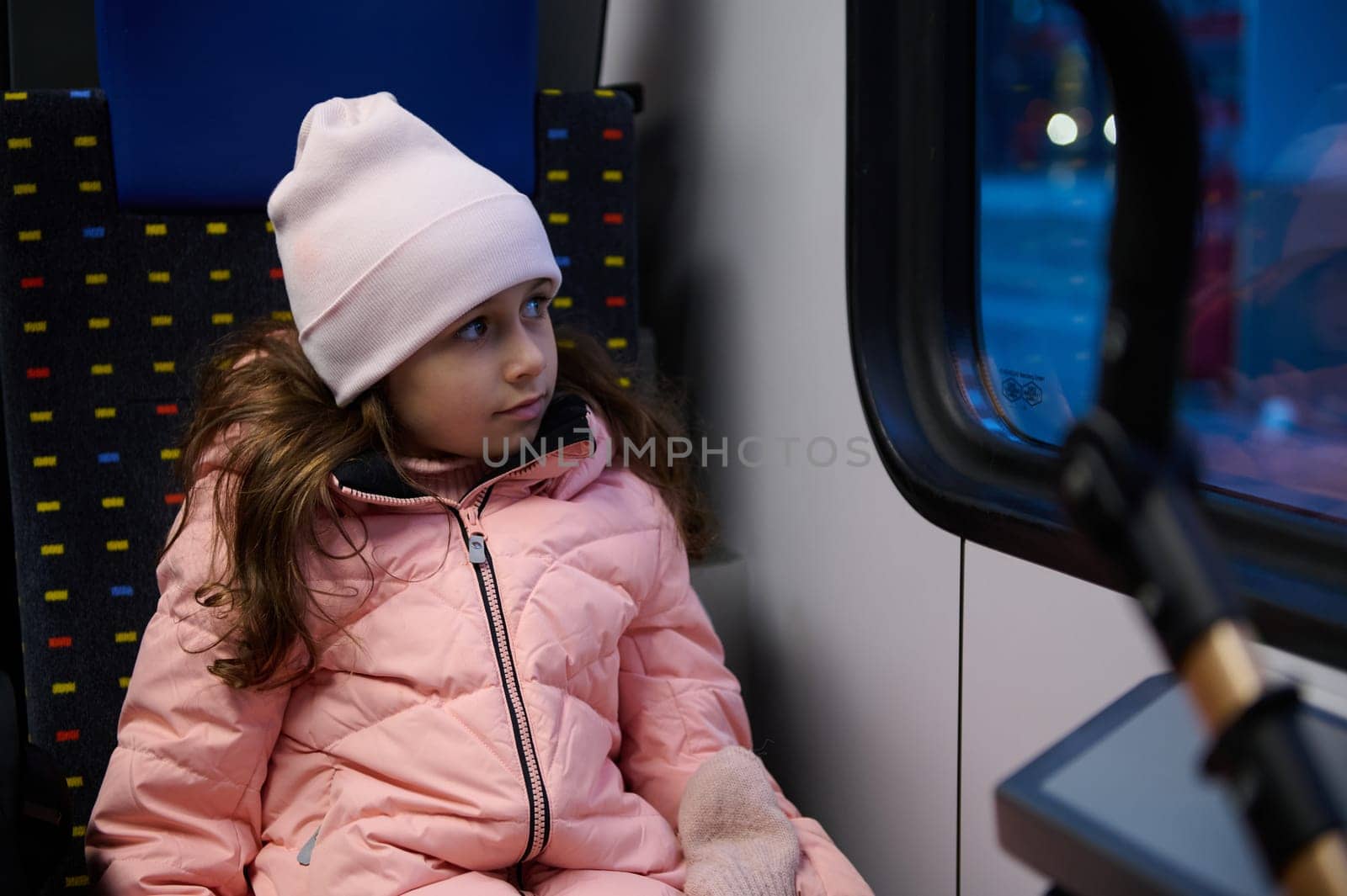 Little child girl in warm winter clothes, enjoys travelling by train, sitting by passenger window, admiring the nature. by artgf