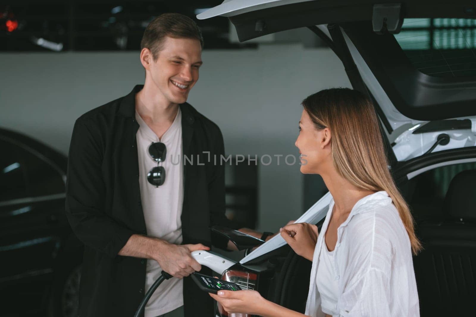 Young couple travel with EV electric car to shopping center parking lot charging in downtown city showing urban sustainability lifestyle by green clean rechargeable energy of electric vehicle innards