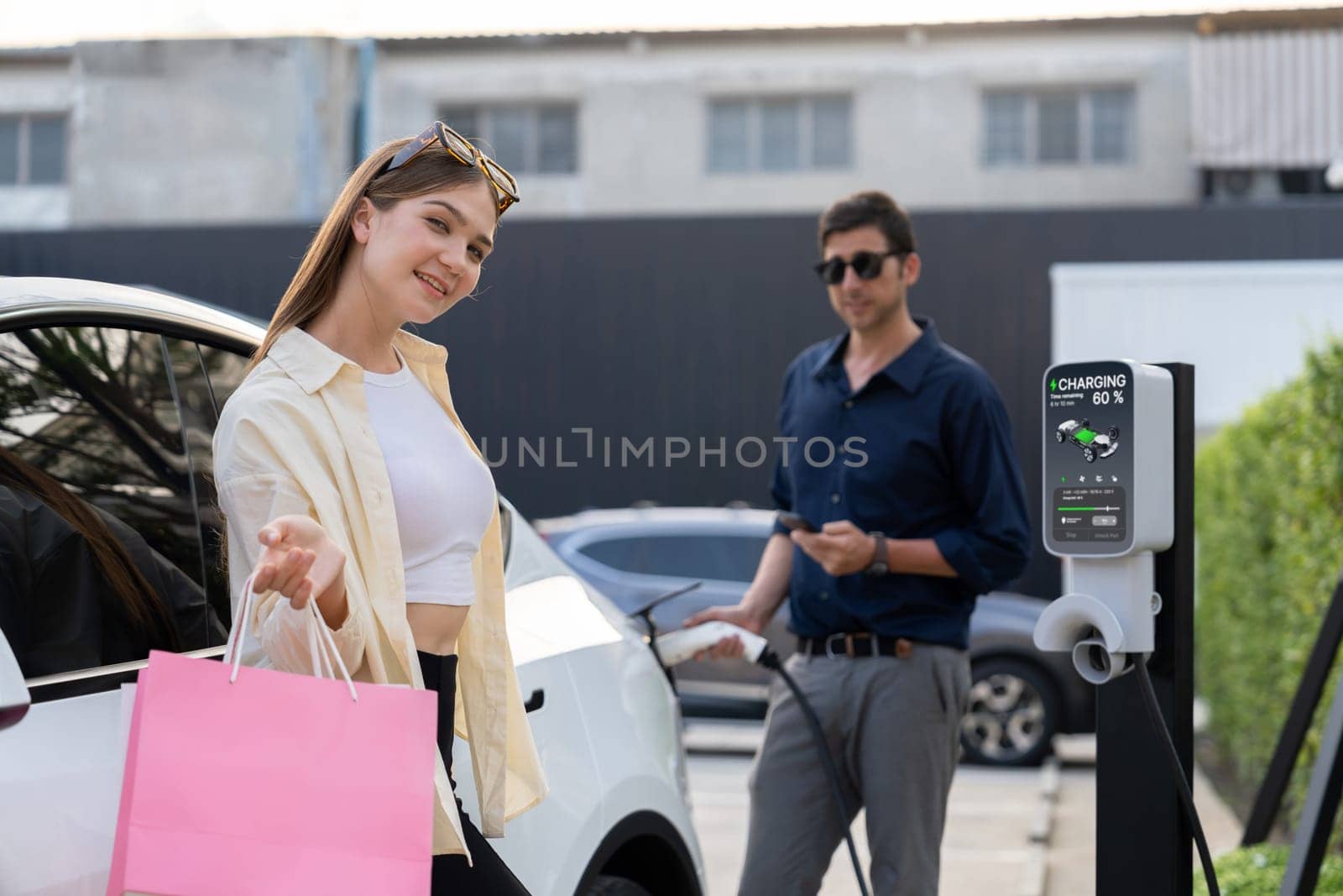 Young couple holding shopping bag and use smartphone to pay for electricity for recharging EV car battery from charging station at city mall parking lot. Happy couple go shopping by eco car. Expedient