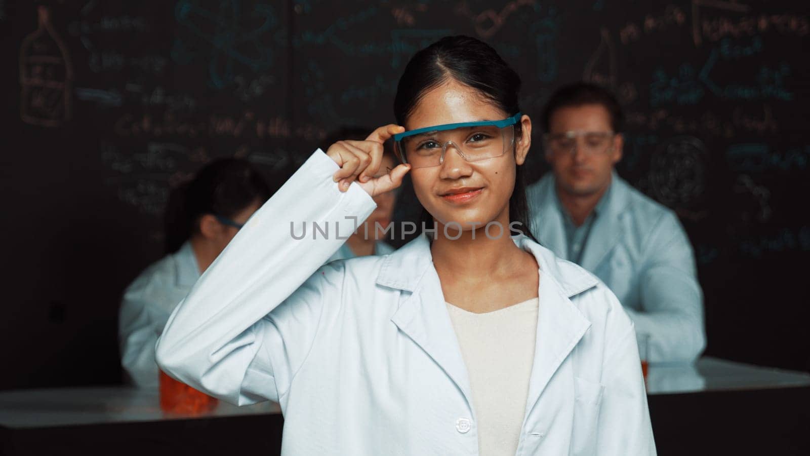 Highschool girl looking at camera while people doing experiment at laboratory. Young cute student pose at camera while standing blackboard with chemical theory with blurring background. Edification.