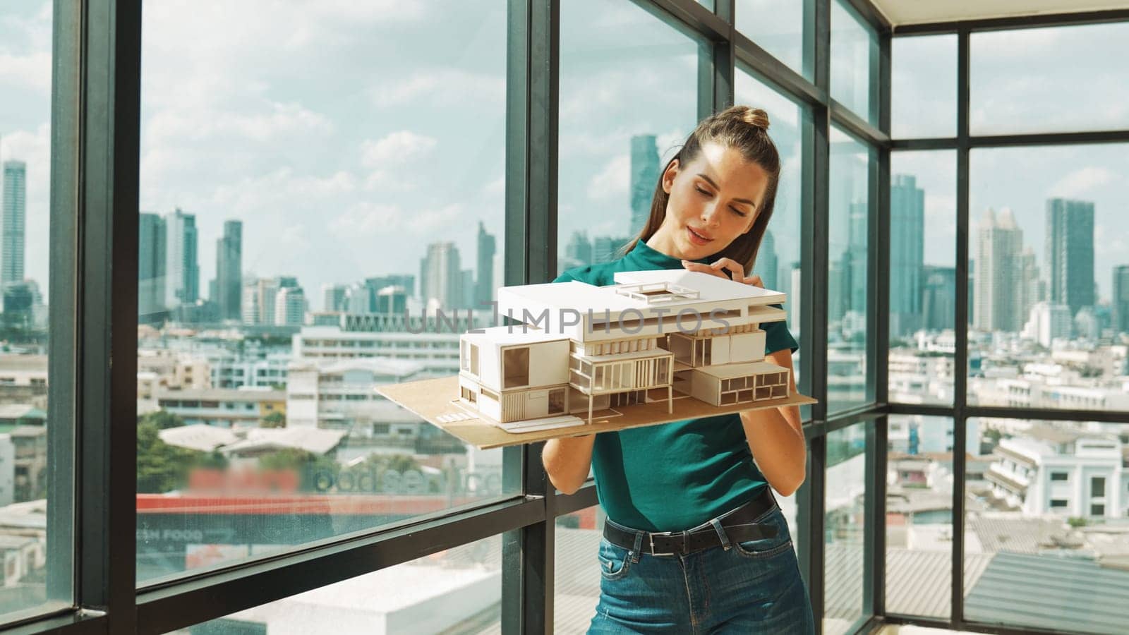 Young smart architect engineer holds architectural model while inspect house model. Professional interior designer checking house construction while standing near window with city view. Tracery
