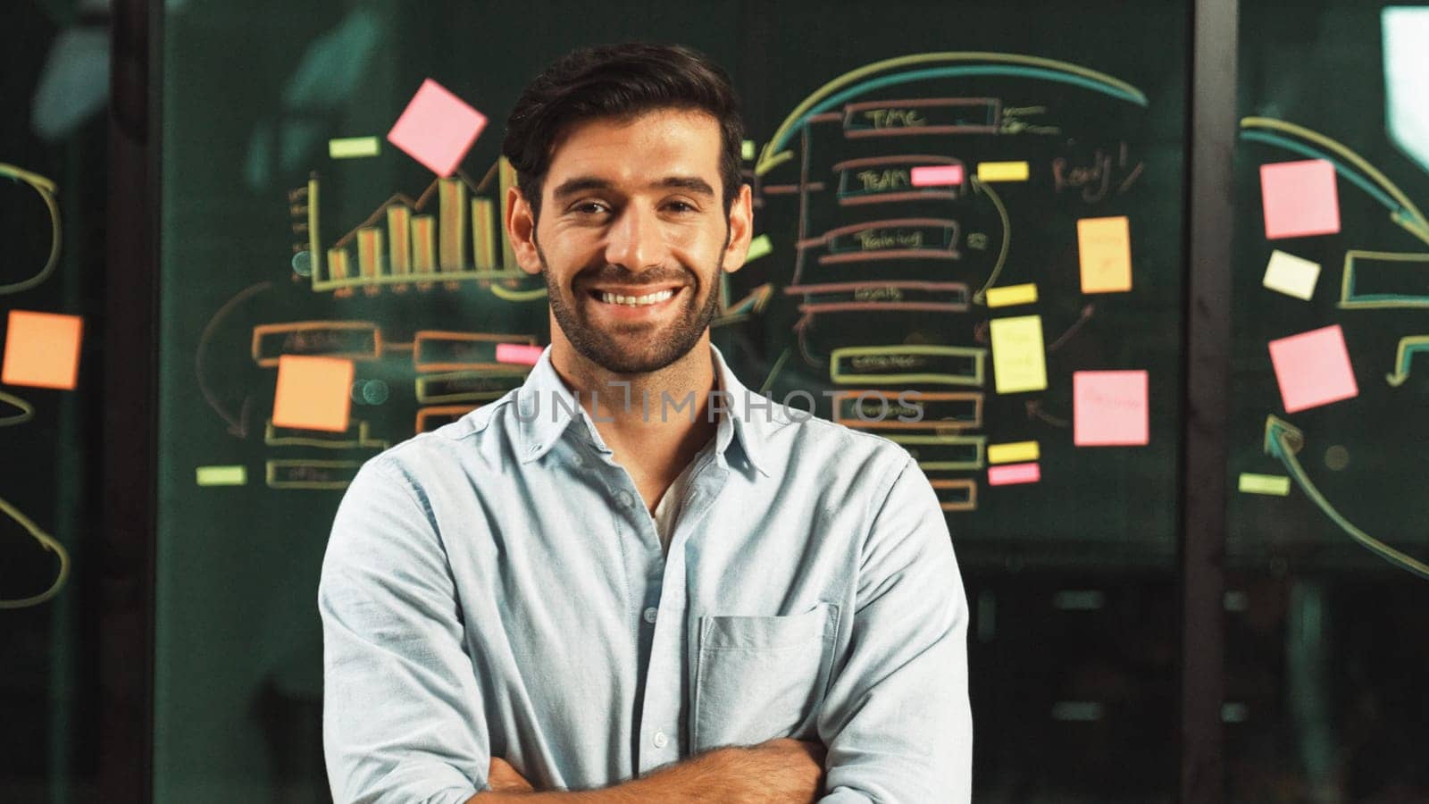 Professional businessman looking at camera while standing with folded arms on glass wall with statistic and sticky notes. Male leader crossing arms while smiling at camera with confident. Tracery