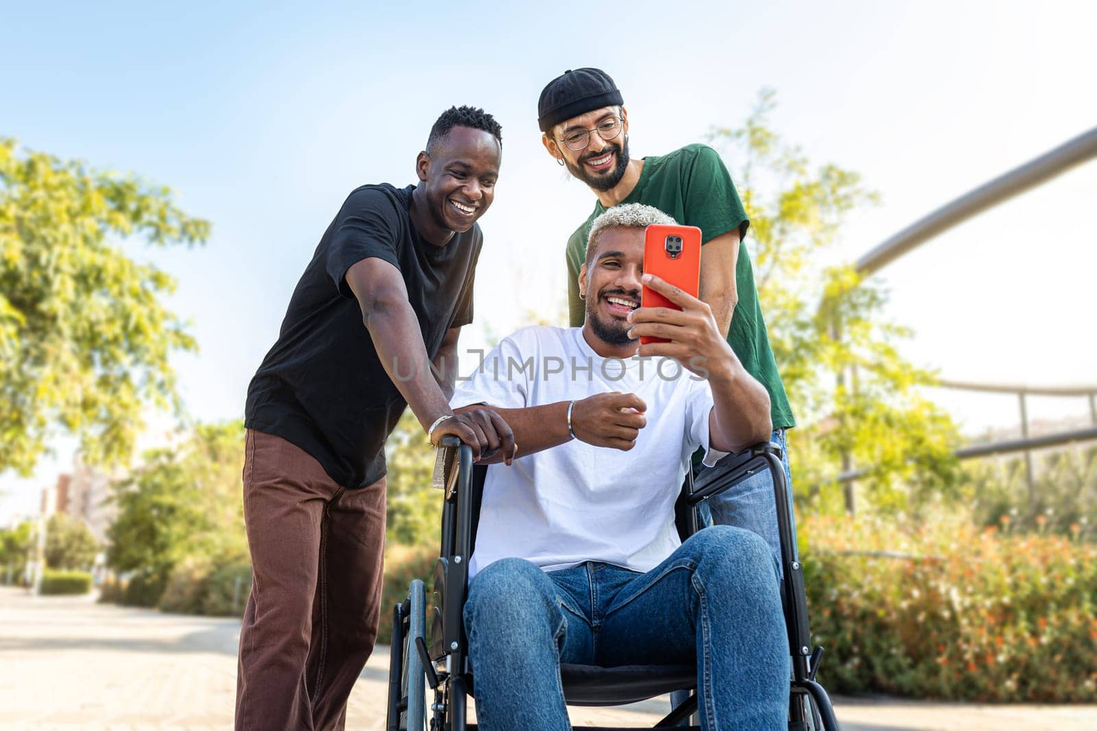 Young African American man in a wheelchair using phone together with male friends outdoors. Friendship and technology.