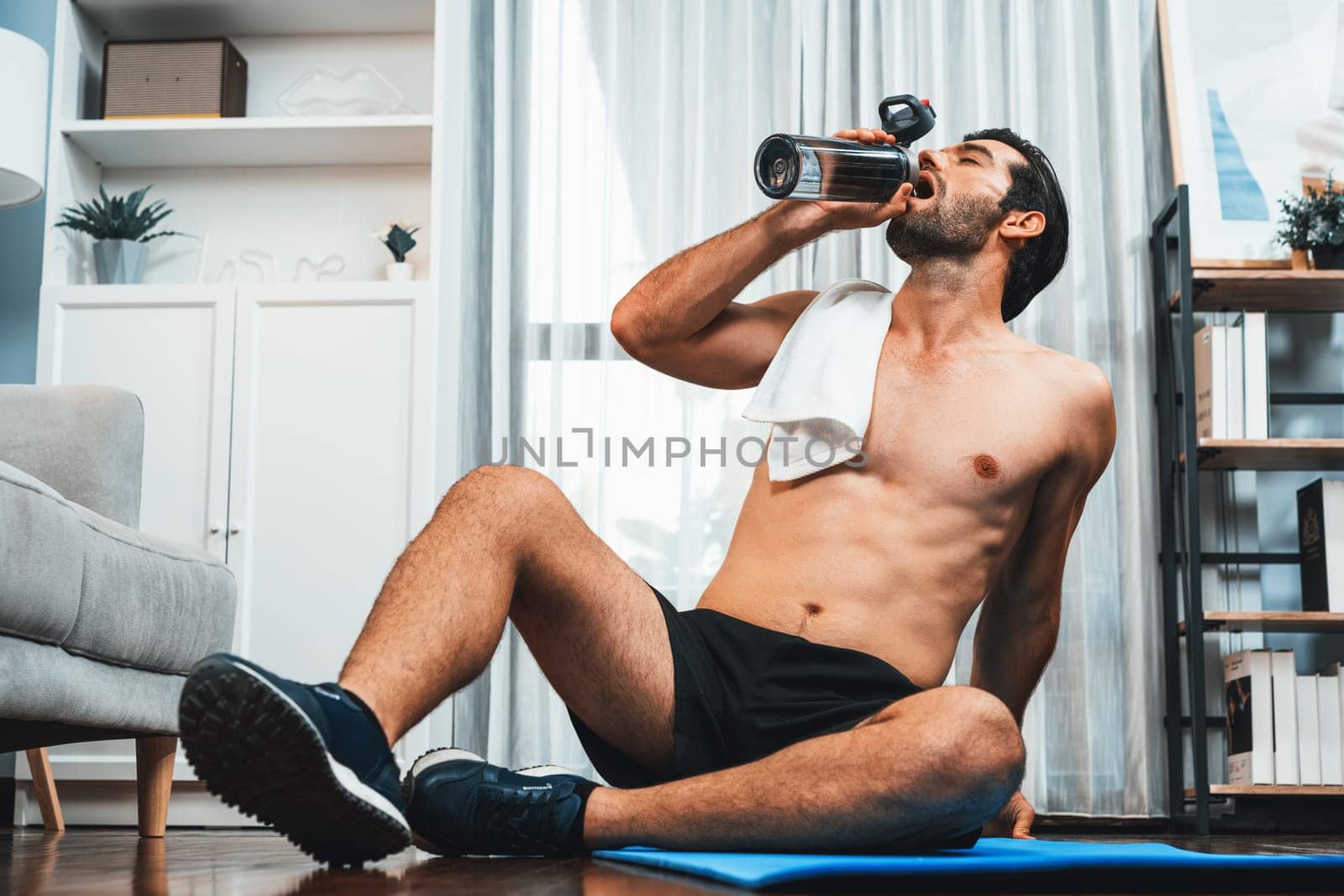 Athletic and sporty man drinking water on fitness mat at gaiety home. by biancoblue