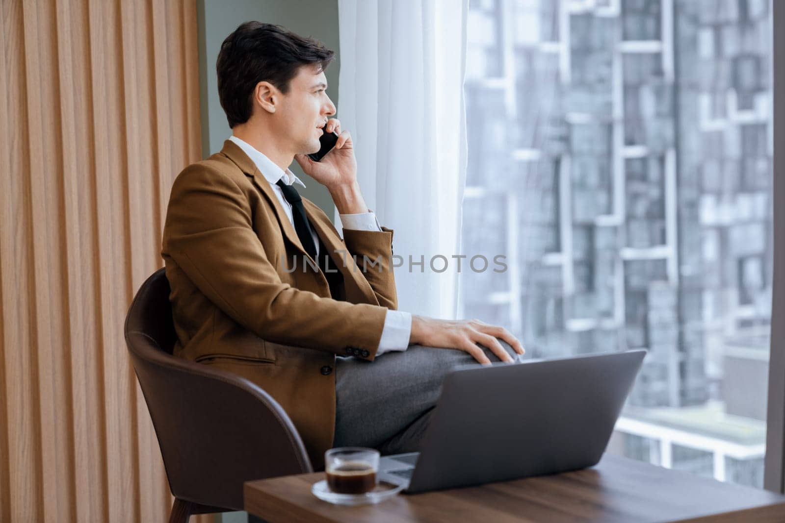 Closeup of handsome businessman making phone call with manager while sitting near window with skyscraper view. Executive manager talking working by using phone and laptop. Look aside. Ornamented.