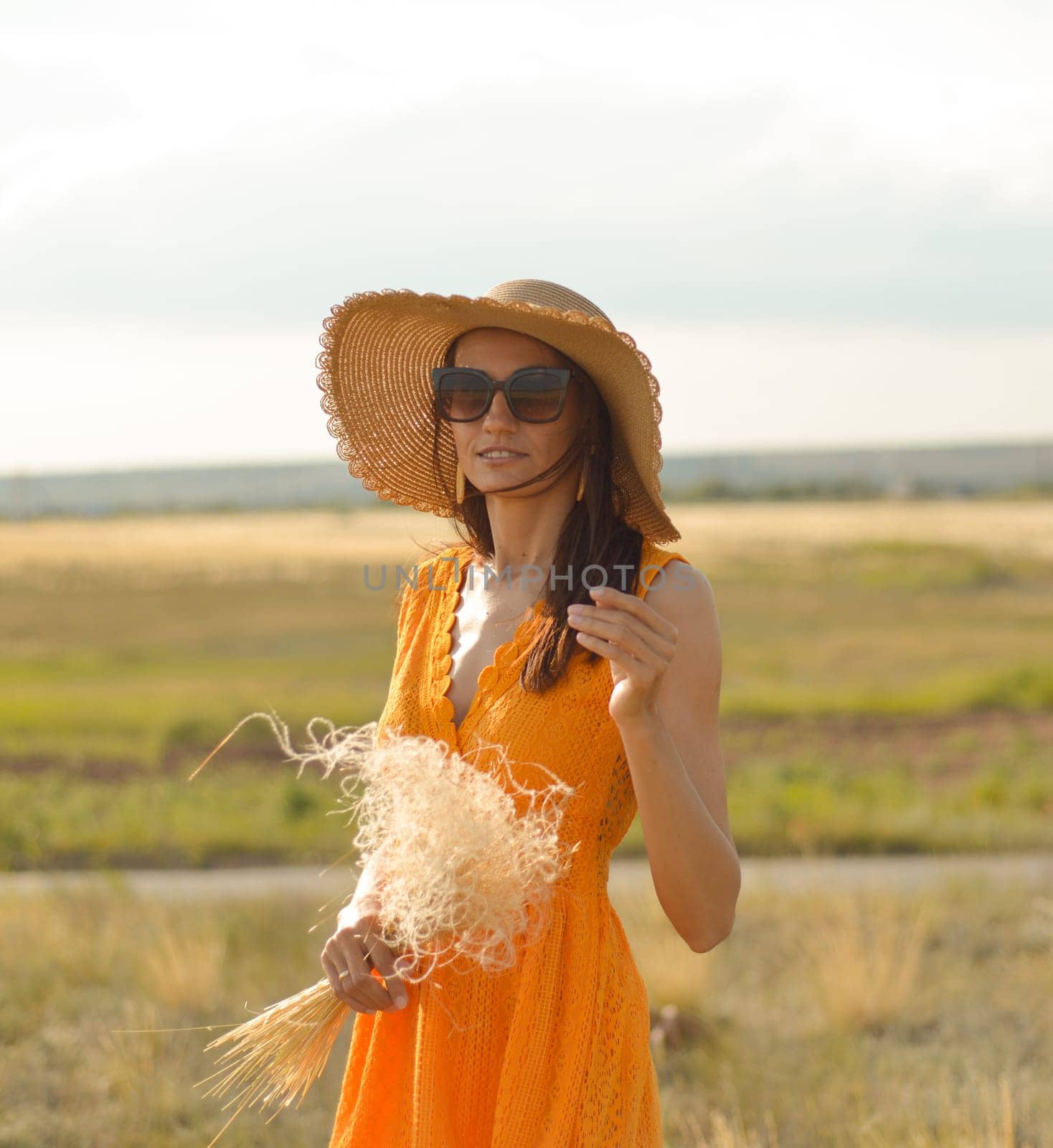 Young woman in an orange dress and a straw hat standing on a field in the rays of the setting sun.