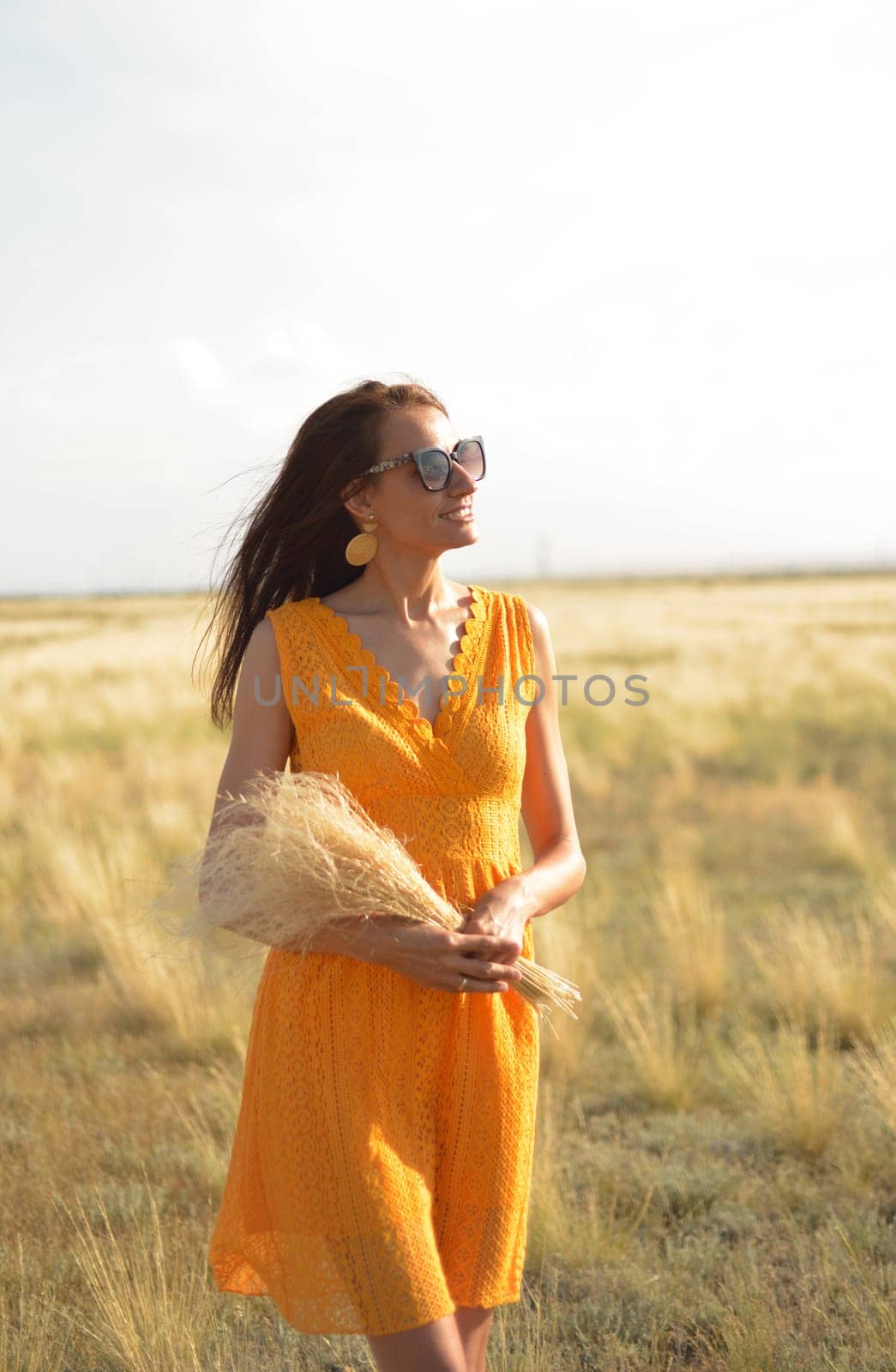 A beautiful brunette girl in an orange dress and sunglasses with a bouquet of field grass enjoys nature in the summer. by Ekaterina34