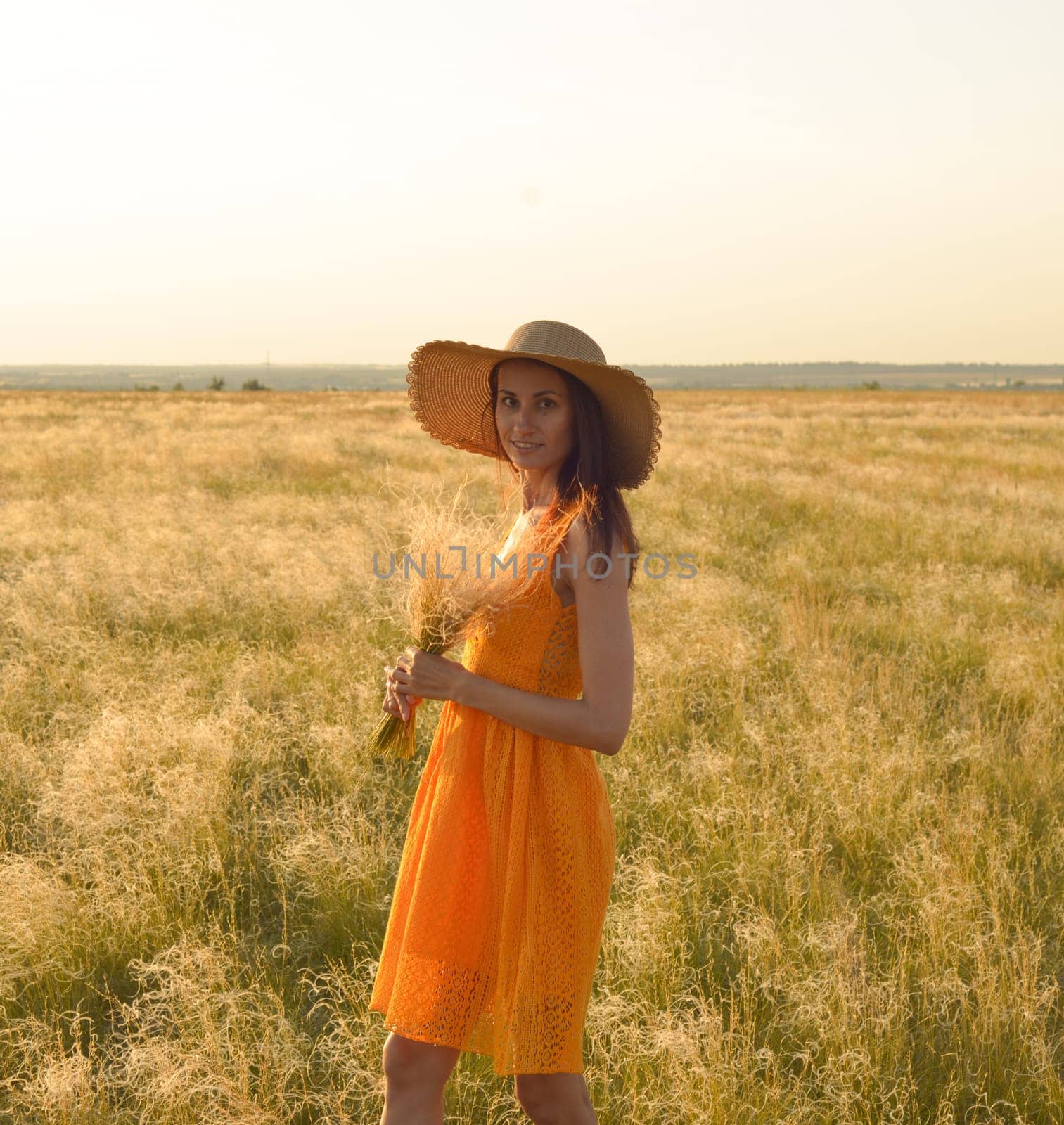 Young woman in an orange dress and a straw hat standing on a field in the rays of the setting sun.