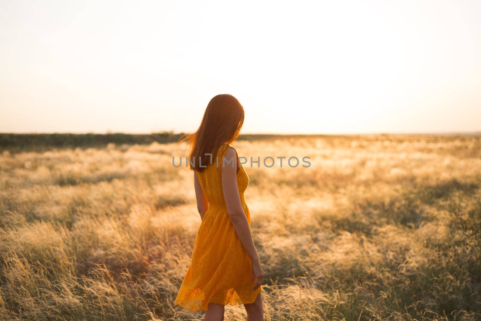 A pretty young girl with long brown hair in an orange dress in a field at sunset. The concept of freedom and nature. by Ekaterina34