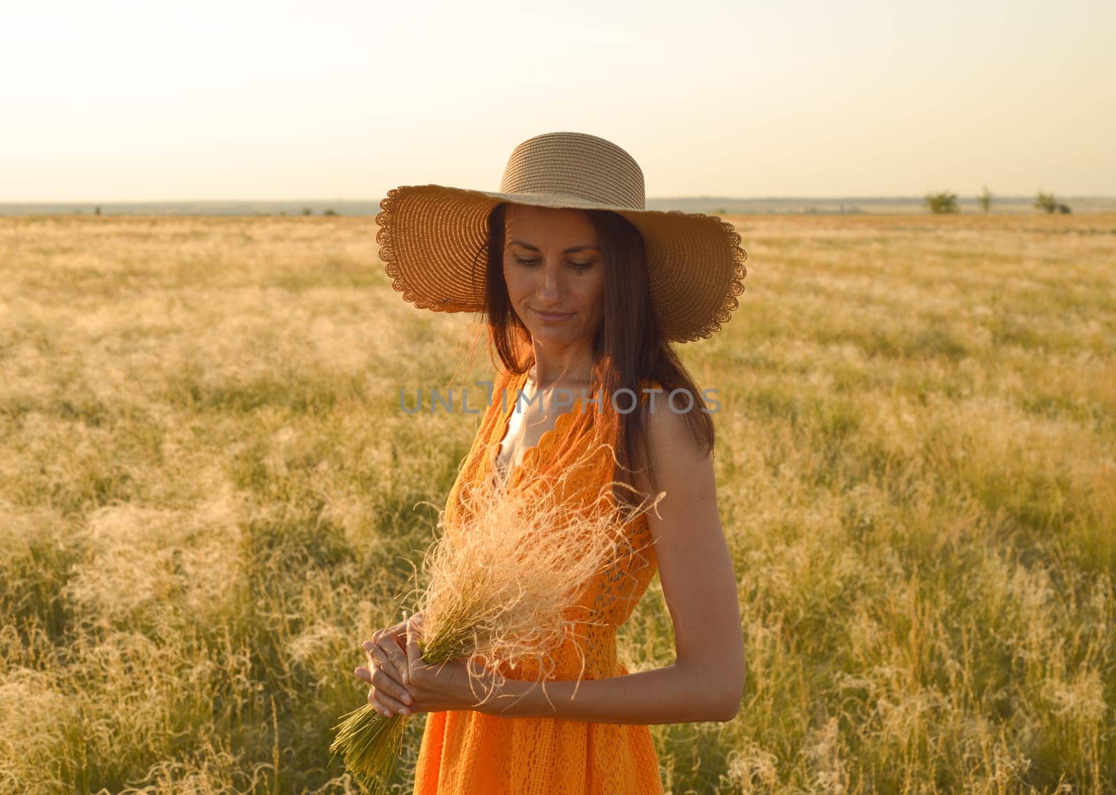 Young woman in an orange dress and a straw hat standing on a field in the rays of the setting sun.