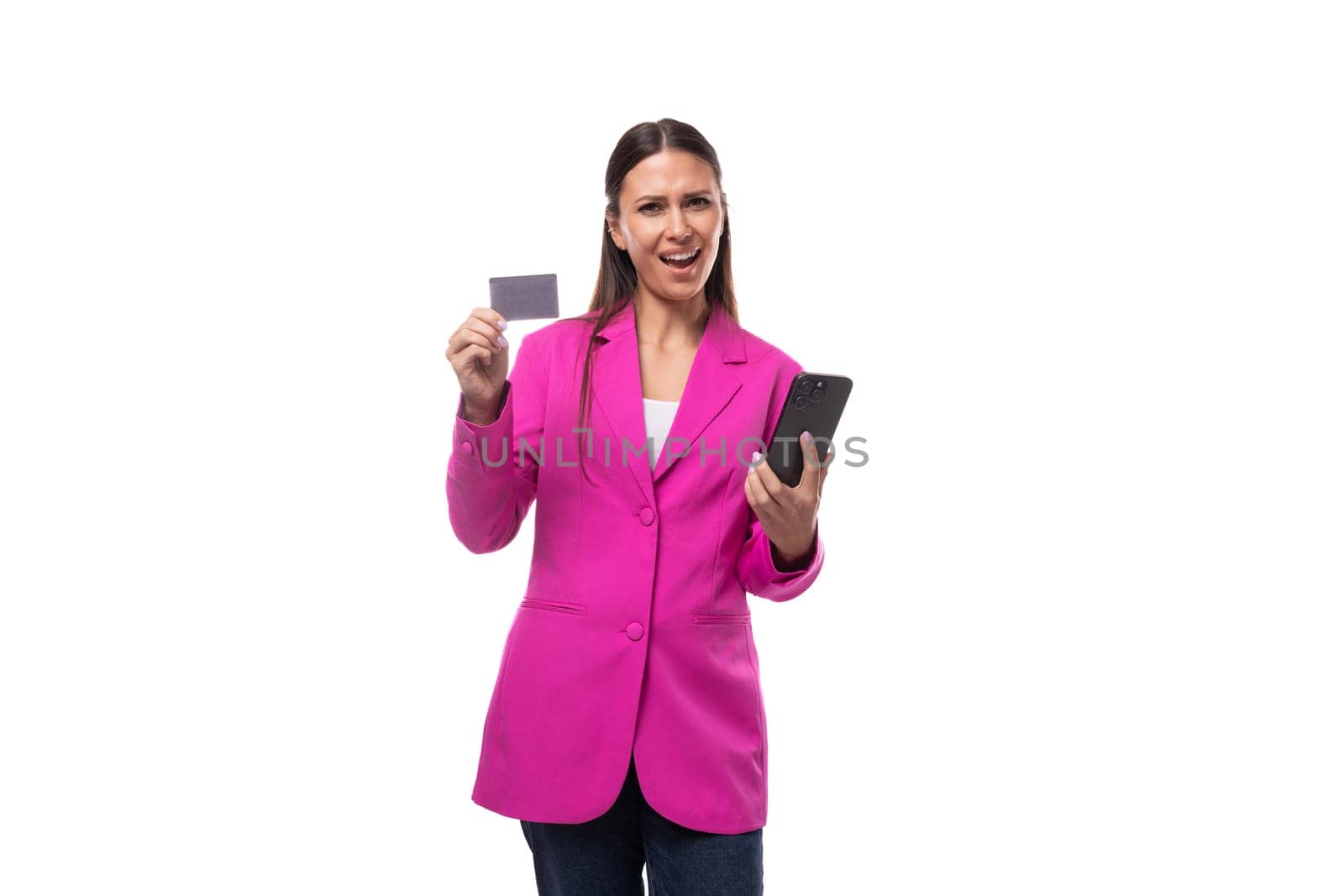 young pretty woman with black straight hair is dressed in a trendy pink jacket holding a credit card and a smartphone.