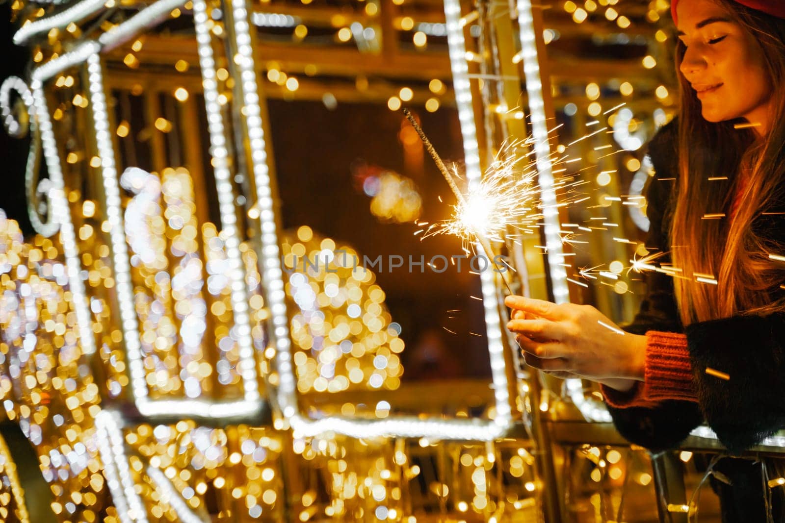 Woman holding sparkler night while celebrating Christmas outside. Dressed in a fur coat and a red headband. Blurred christmas decorations in the background. Selective focus.