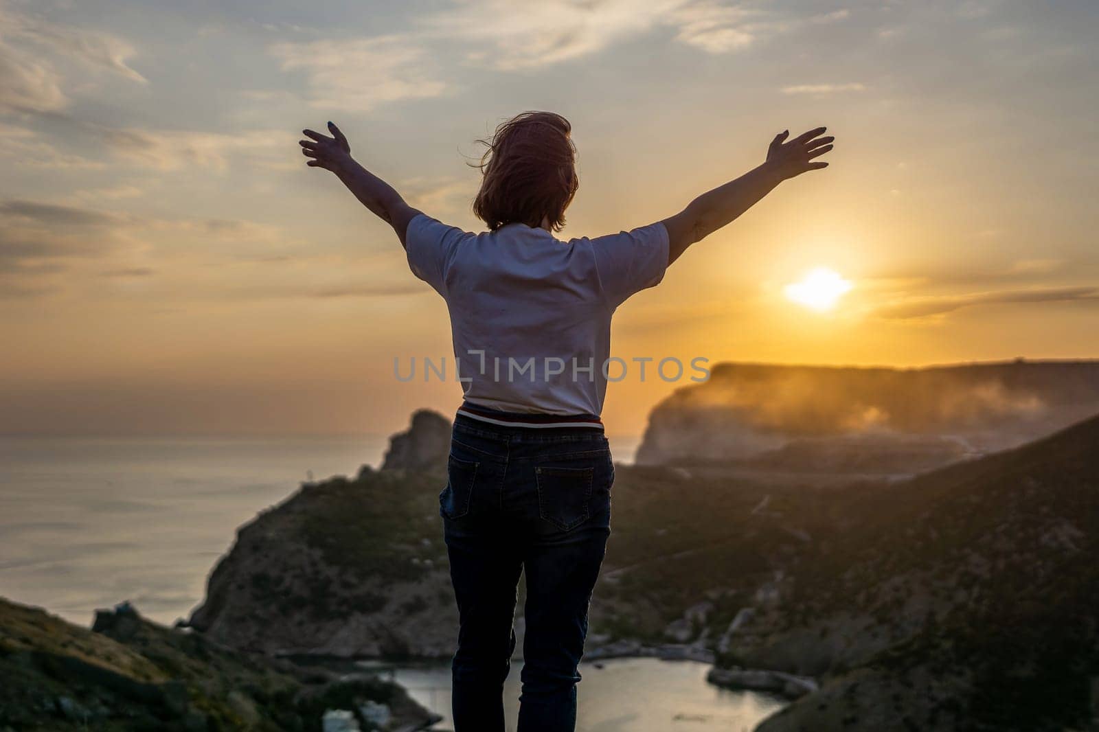 Happy woman on sunset in mountains. Woman standing with her back on the sunset in nature in summer with open hands. Silhouette. by Matiunina