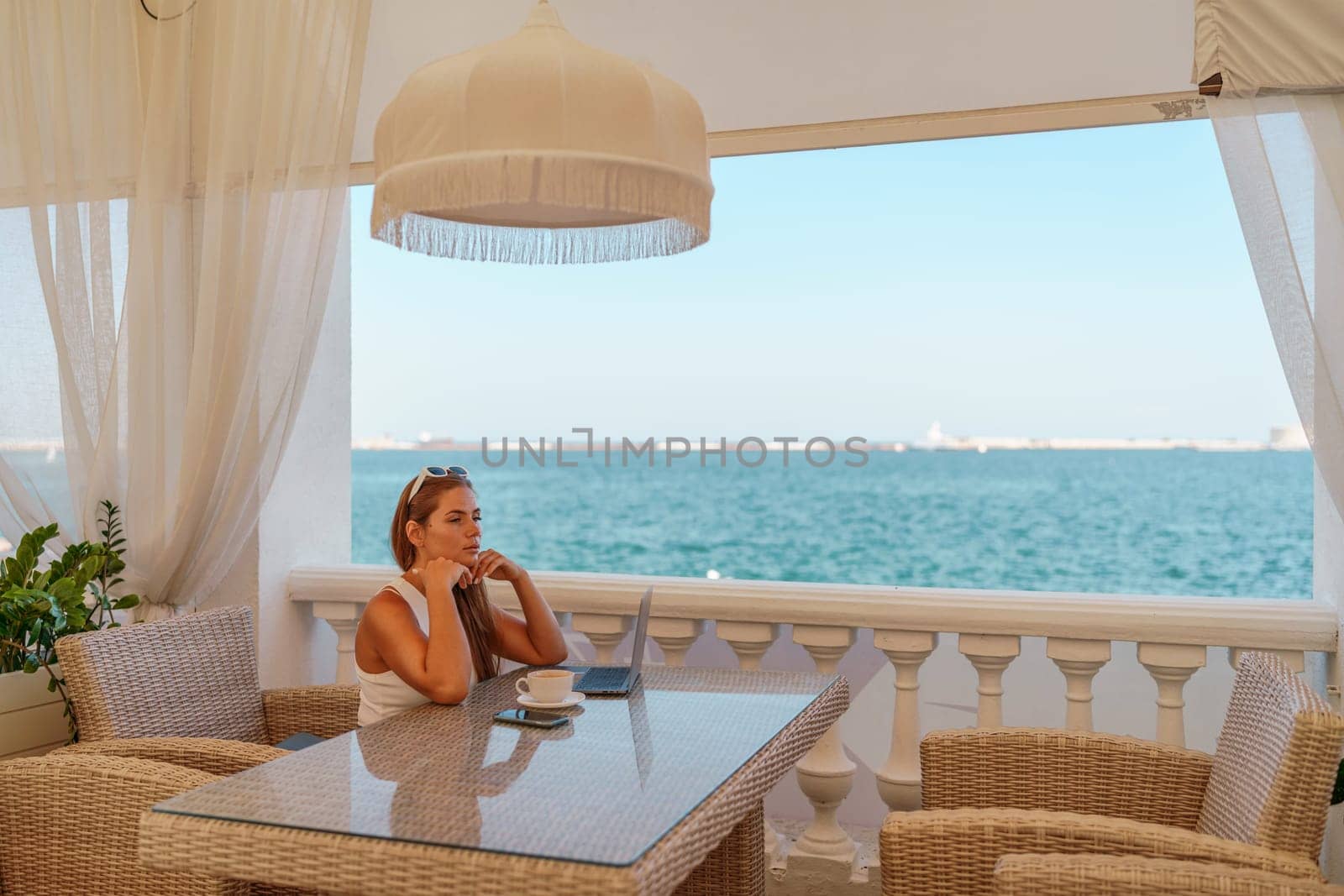 Woman coffee cafe laptop sea. Female freelancer using laptop on coffee break in modern cafe with sea view in the morning by Matiunina